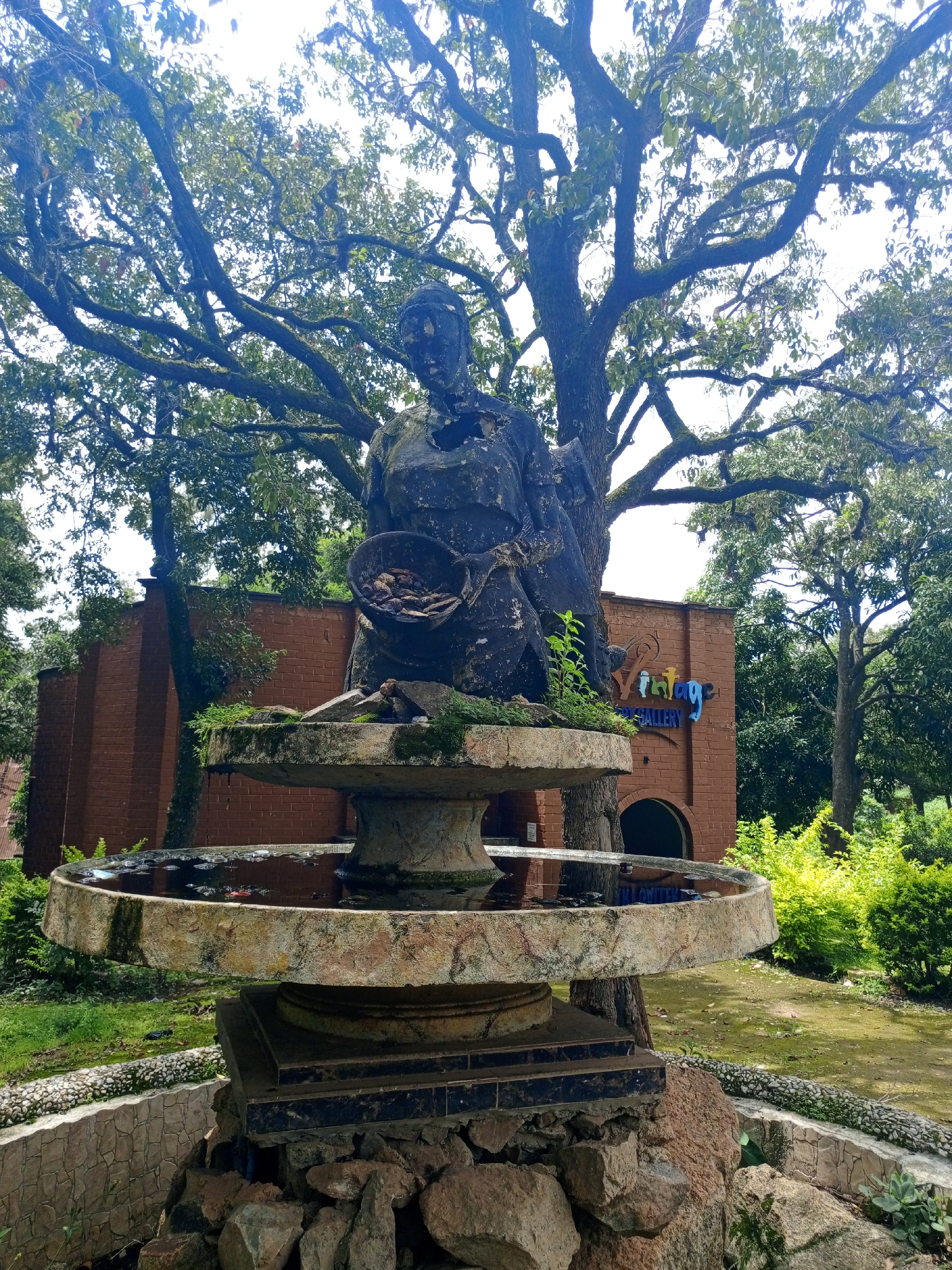 This image shows a stone fountain with a statue of a person holding a bowl, in front of a brick building with a sign that reads "Vintage Art Gallery". The statue is weathered and appears to be old. The fountain is filled with water, and there are some plants growing around it. The building behind the fountain is a single-story structure with a curved entrance. The area is surrounded by trees and greenery. This scene is located in Jos, Nigeria, at the Vintage Art Gallery.