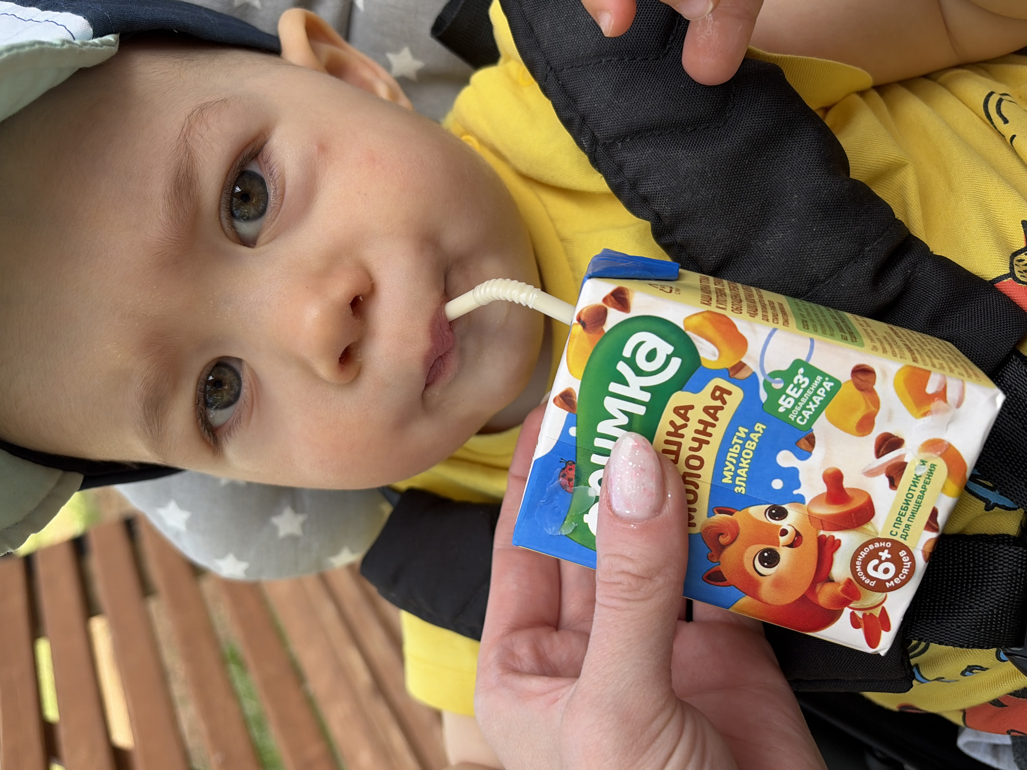 A baby is drinking from a juice box. The juice box is labeled "Агуша Молочная Мульти Злаковая" and "Без Добавления Caxapa". The juice box is held by a person with manicured nails. The baby is wearing a yellow shirt and a hat. The scene is set in Chaikovsky, Russia, based on the juice box brand.