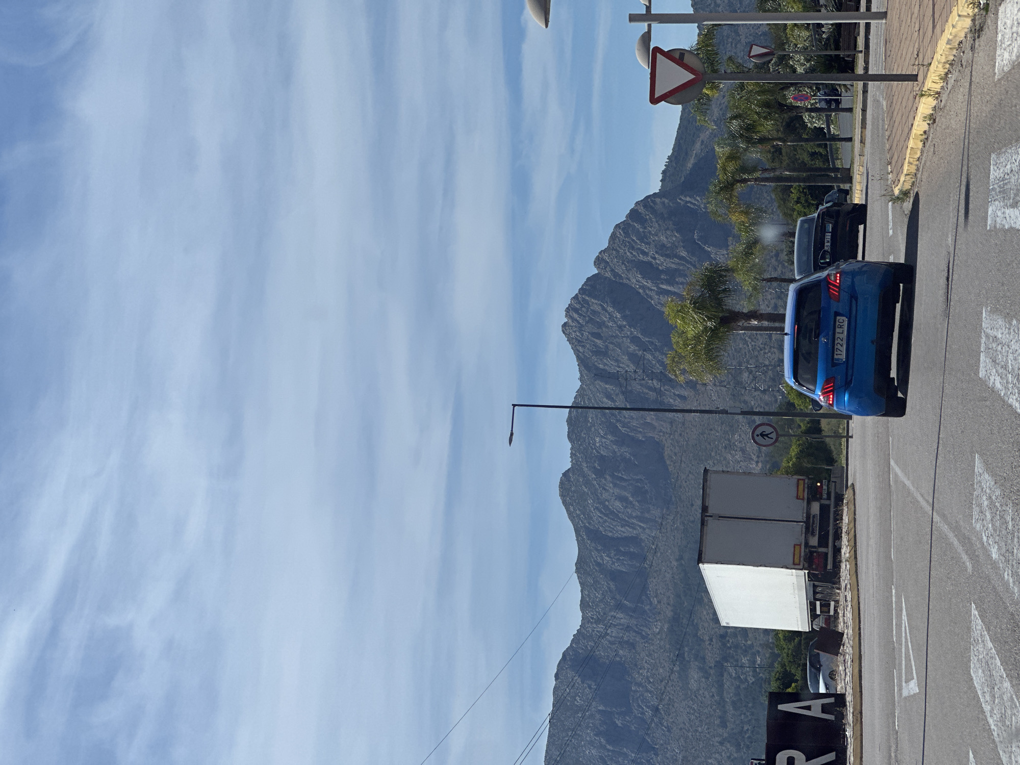 The image shows a street in Ondara, Spain, with a mountainous backdrop and a clear sky with scattered clouds. A blue car with license plate "1222 LRC" is parked on the right side of the road, followed by a gray vehicle. A white truck is further down the road. Traffic signs are visible along the roadside. The road has zebra crossings. The landscape includes palm trees and other greenery. The word "ONRA" can be seen on a sign.