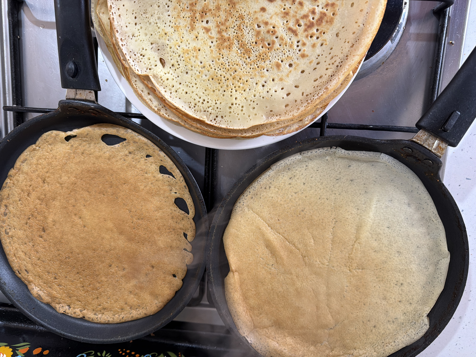 The image captures a culinary scene in a kitchen, likely in Chaikovsky, Russia, focusing on the preparation of pancakes or blini.
The main subjects are a stack of finished pancakes on a white plate, and two additional pancakes cooking in separate black frying pans. The white plate, laden with golden-brown pancakes, is positioned centrally over a metallic stovetop, with portions of it extending over two burner grates. These stacked pancakes display a classic porous texture, indicating they are freshly made.
Below the plate, two black non-stick frying pans are visible, each containing a pancake in different stages of cooking. The pancake in the left pan is uniformly golden-brown, suggesting it is nearly or fully cooked. The pancake in the right pan is lighter in color, appearing to be in an earlier stage of cooking, with a faint wisp of steam rising from its surface. Both pans have black handles and show signs of use, such as wear on the metal parts connecting the handles to the pan body and some residue on the pan edges.
The setting is a home kitchen, with a metallic gray stovetop and black grates visible beneath the pans. A white countertop is visible on the far right. The activity taking place is clearly the cooking of pancakes, showcasing both completed items and those in progress.
There are no people visible, nor any discernible text. The time of day, weather, and specific emotions cannot be determined from the image, but the scene conveys a sense of domestic food preparation.
