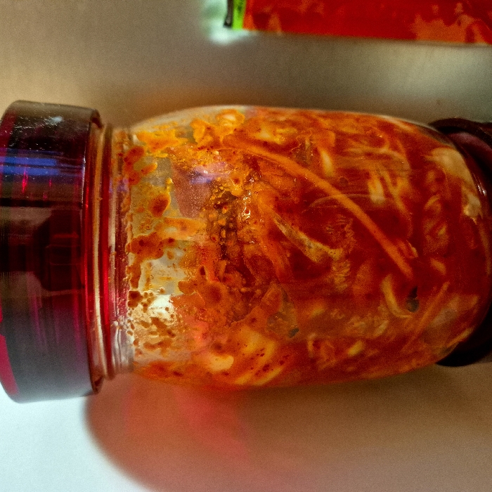 A glass jar sits horizontally, filled with a vibrant red and orange mixture of pickled vegetables. The contents appear chunky and stringy, suspended in a thick, oily sauce. The jar has a dark red lid, which is screwed on tight. Light reflects off the glass, creating a sheen across the surface. A shadow is cast below the jar, suggesting a bright light source from above. A partially visible red and green object sits in the background.
