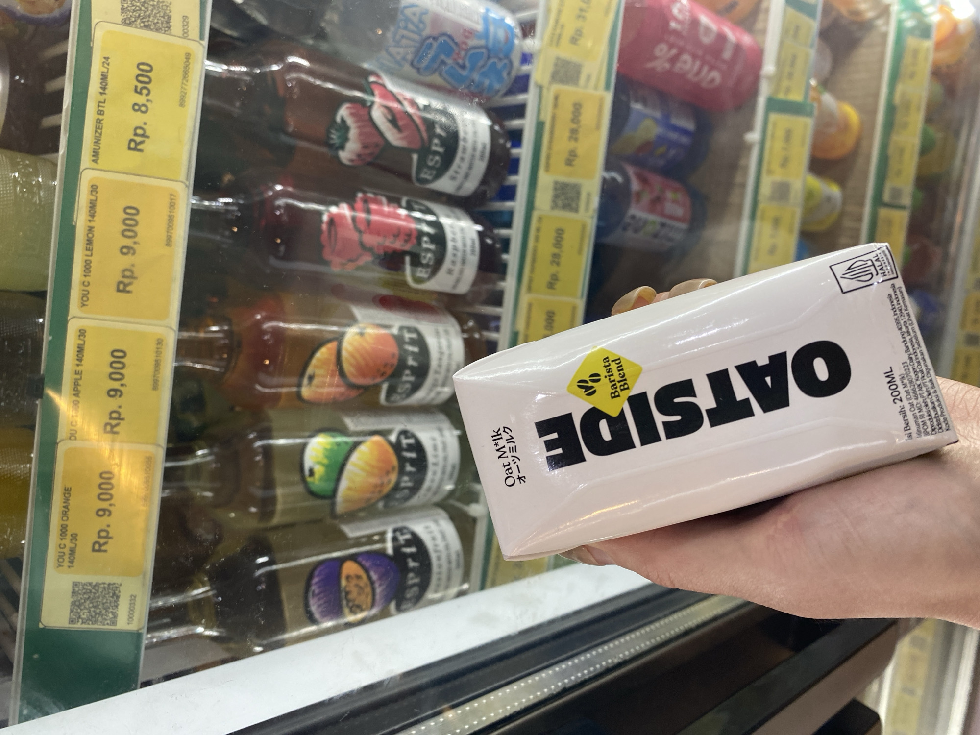 A person is holding a carton of "Oatside Barista Blend Oat Milk" in front of a refrigerated display case at a store in Kuta Selatan, Indonesia. The Oat Milk carton is white with black lettering. The refrigerated display case contains rows of glass bottles with colorful fruit designs on their labels, like Esprit brand. Yellow price tags are affixed to the refrigerator shelf in front of the bottles, displaying prices in Rupiah (Rp.). These prices include Rp. 8,500 for Amunizer, Rp. 9,000 for You C 1000 Orange, Apple and Lemon flavor. Other tags with higher prices (Rp. 28,000 and Rp. 31,000) are also partially visible. The refrigerator light illuminates the products. The hand holding the milk carton is fair-skinned, and its nails are light-colored. The context suggests a shopping environment.
