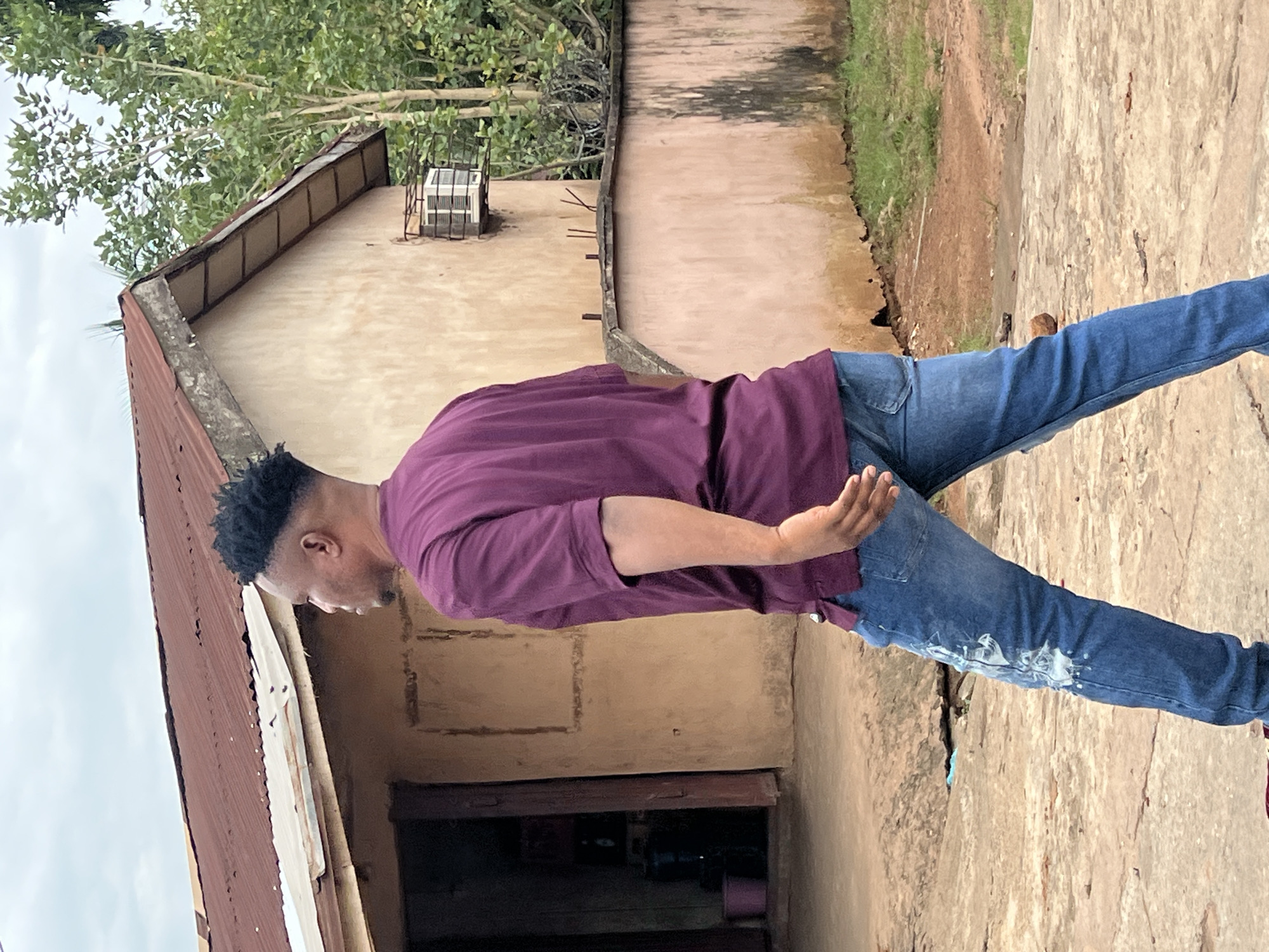 A man with a fade haircut, wearing a maroon t-shirt and blue jeans, walks away from the camera in front of a single-story, light-brown building with a corrugated metal roof. The building appears to be in disrepair, with a weathered facade and exposed beams under the eaves. An air conditioning unit is mounted on the exterior wall, protected by a metal grate. A pink bucket is visible near the entrance of the building, and a dark blue water dispenser is inside. A concrete wall with barbed wire runs along the side of the property, bordering an area with grass and trees. The ground in the foreground is a rough, uneven concrete surface. The scene is set outdoors with overcast skies. The location is Nkpor, Nigeria.