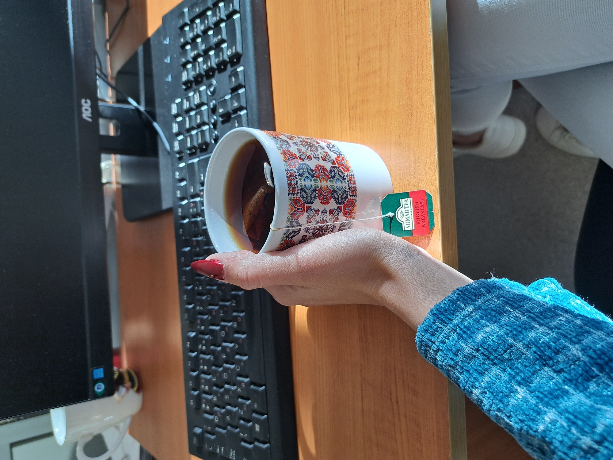 A woman is holding a cup of tea with an "Ahmad Tea English Breakfast" tea bag inside. The cup has a patterned design around the exterior. She is wearing a blue sweater and has red nail polish. The scene is in an office, as a black computer monitor and keyboard are visible on a wooden desk. The specific location in Sofia, Bulgaria cannot be determined from the image.