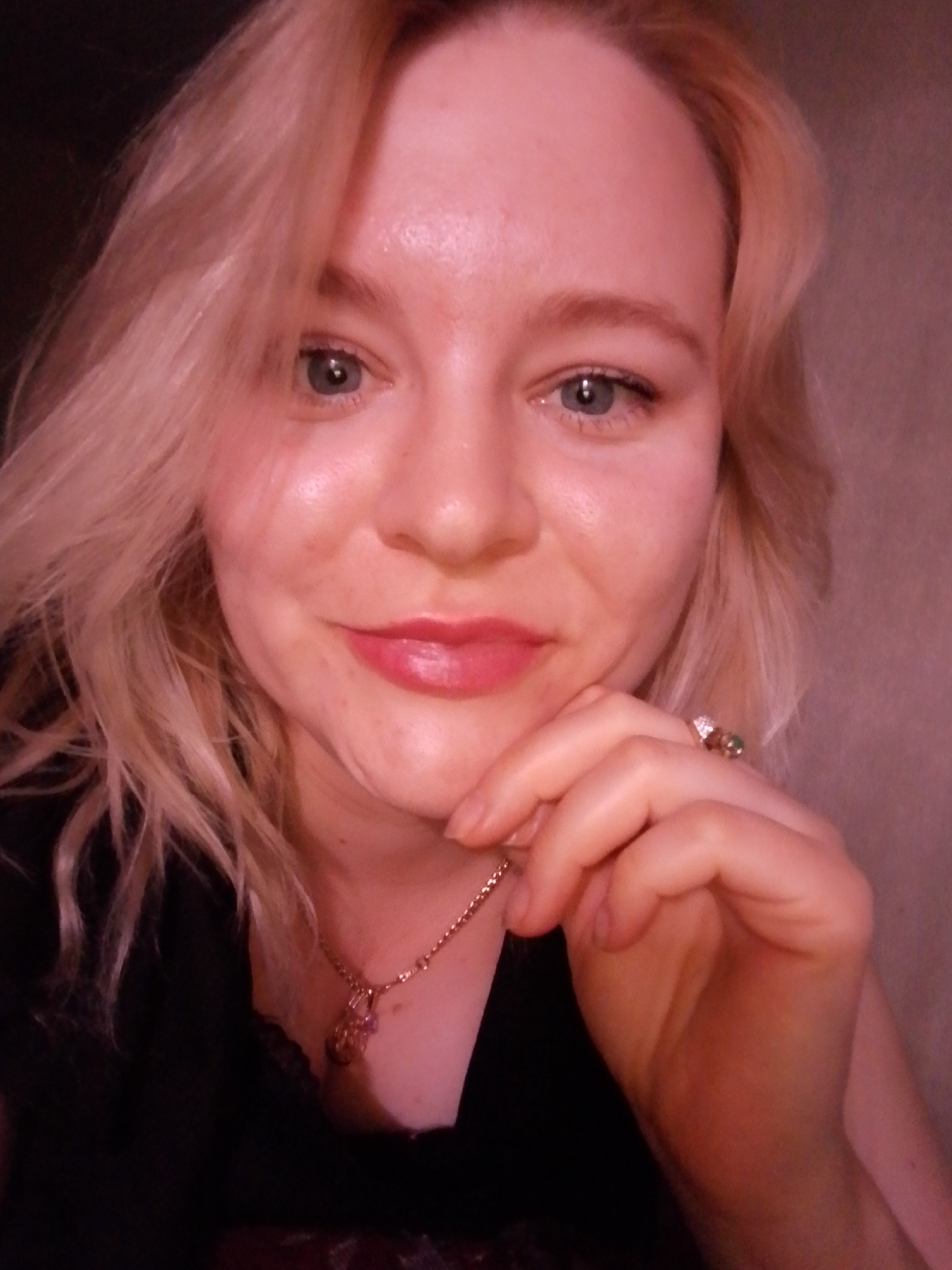 The image depicts a close-up, head-and-shoulders portrait of an adult female, reportedly located in Izhevsk, Russia. The subject has blonde hair, light-colored eyes, and fair skin, which appears flushed with a reddish hue under soft, warm lighting. She is looking directly at the camera with a slight smile, revealing a trace of light-colored lipstick on her lips. Her right hand is raised, with fingers resting gently against her chin and lower jawline. She is wearing a dark-colored top, possibly black, with visible lace trim near the neckline. Around her neck is a delicate gold-toned chain necklace with a small, light-colored pendant featuring a pink or purple stone. A ring with two prominent stones, one green and one lighter, is visible on the ring finger of her right hand. The background is an indistinct, solid-colored surface, likely a wall, appearing in shades of pinkish-brown due to the ambient lighting, and lacks any distinguishing features or objects.