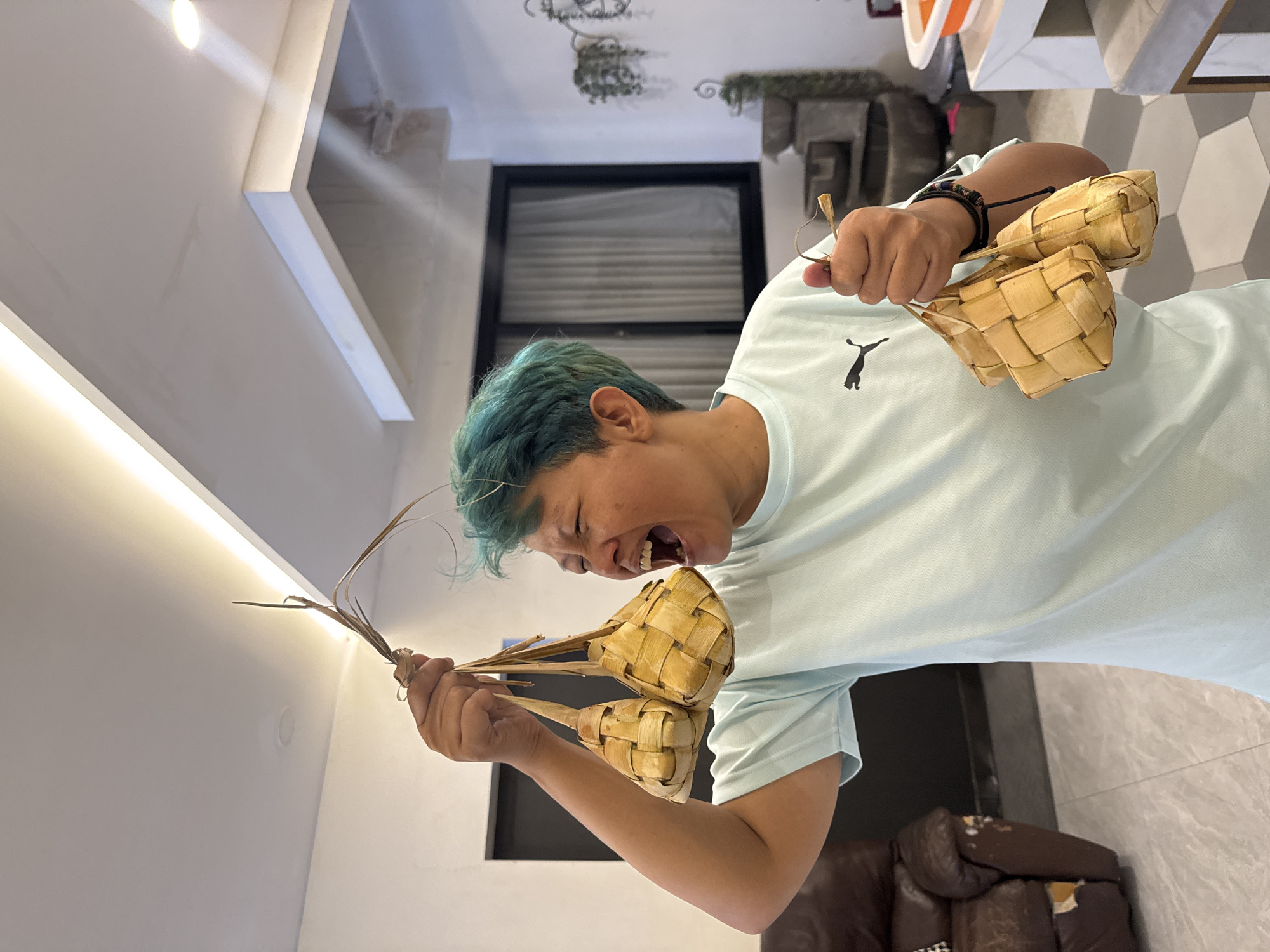 A person with blue-green hair is holding up several ketupat in an indoor setting. The individual is wearing a light blue t-shirt with a black Puma logo. The ketupat are traditional rice cakes wrapped in woven palm leaves. The background shows elements of a modern interior with a dark framed window and a tiled floor. A brown leather chair is partially visible in the foreground. This is likely a residential setting in Bekasi, Indonesia.