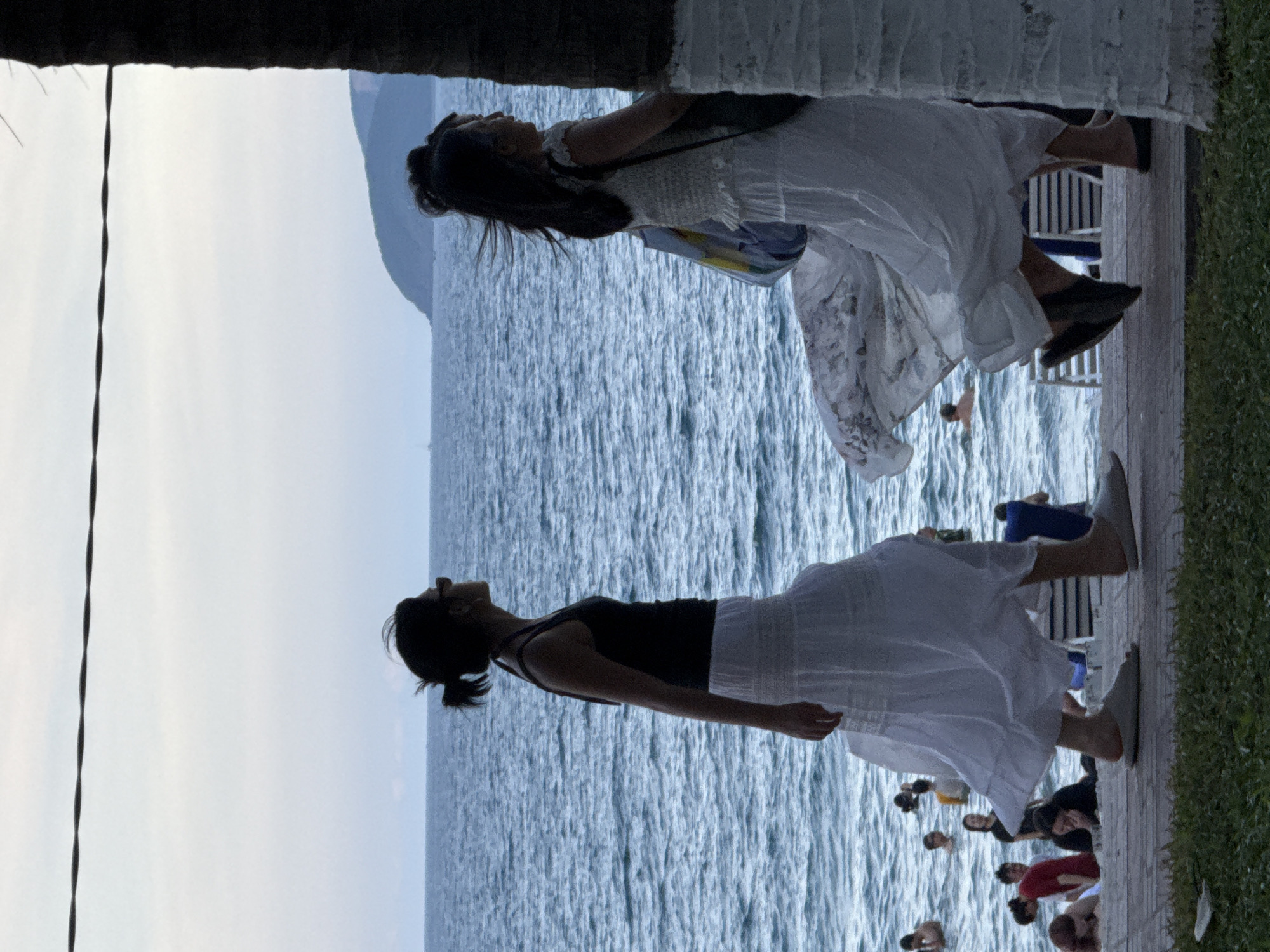 This media file, captured in Nha Trang Ward, Vietnam, depicts a group of people by the sea, likely during a gathering or event. The scene is oriented vertically, with the sea occupying the majority of the frame. The water appears calm and blue, with small ripples suggesting a gentle breeze.
In the foreground, two women are prominent. One is silhouetted against the bright sky, wearing a white, flowing dress and carrying a bag. Her head is tilted back, and her arms are outstretched, suggesting a moment of freedom or contemplation. The other woman, also in silhouette, wears a black tank top and a white skirt. She is leaning back with her head tilted upwards, appearing relaxed.
Further into the background, other individuals are visible near the water and on a wooden boardwalk. Some are seated, while others appear to be standing or walking. The presence of beach chairs and umbrellas hints at a public area like a beach or resort.
The overall mood is serene and peaceful, with the soft light suggesting either early morning or late afternoon. The vibrant blue of the sea and the lush green grass on the right side of the frame add to the tropical atmosphere. There is no visible text in the media file.