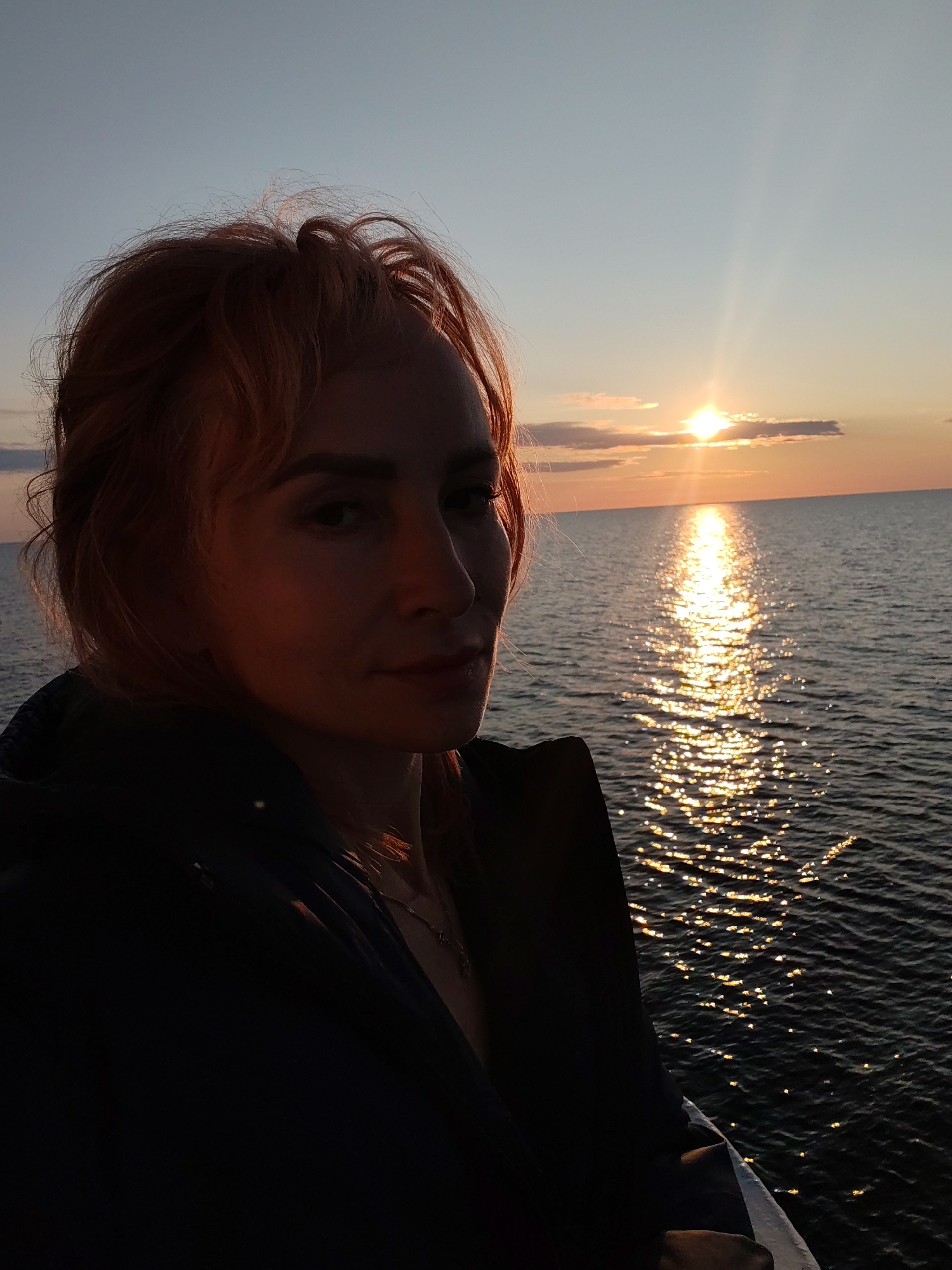 A medium shot photograph captures a woman with reddish-blonde hair looking towards the camera. She is positioned on the left side of the frame, with her face partially illuminated by the setting sun. Behind her, a vast body of water stretches to the horizon, reflecting the warm golden hues of the sunset. The sun itself is a bright, central point of light in the upper right quadrant of the image. Wispy clouds are visible in the sky, tinged with orange and pink. The woman is wearing a dark jacket. The scene appears to be an outdoor setting, possibly on a boat or a pier, overlooking the sea at dusk. The location is specified as Kakkarovo, Russia.