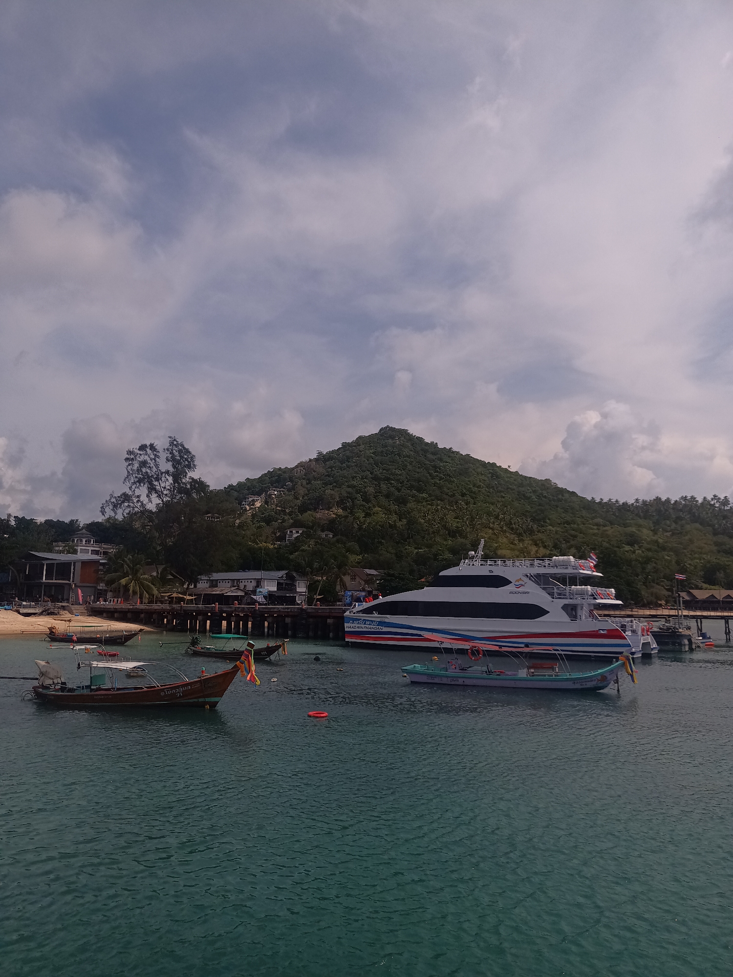 The image depicts a coastal scene in Tambon Ko Tao, Thailand. In the foreground, the water is a turquoise-green color. Several small traditional Thai fishing boats are docked in the water. One boat, made of wood, has a number "71" painted on it along with Thai script and colorful streamers hanging from the back.
In the background, a larger modern ferry is docked near a wooden pier, a white and blue ferry with "BOONSIRI" written on its side. The pier is connected to the shore, which has a beach with buildings behind it. A large, green, mountainous hill is visible behind the buildings and pier, and a cloudy sky occupies the top part of the frame.