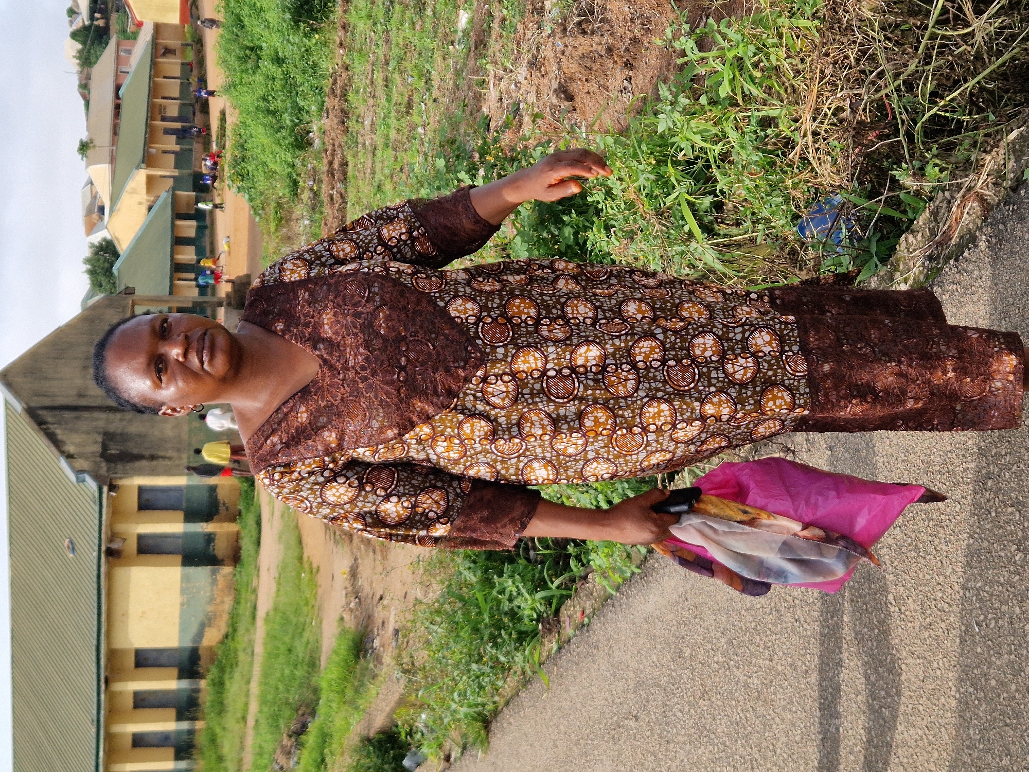 A woman stands on a paved path in front of a school in Kabong, Nigeria. The woman is wearing a brown and white patterned dress with brown lace trim. She is holding a pink plastic bag in her left hand containing a few folded pieces of fabric. The school building behind her is painted yellow and green. In the background, other school buildings and a hilly landscape are visible. Several children are playing in a courtyard in front of one of the buildings. The ground is a mix of dirt and overgrown grass. The sky is overcast.