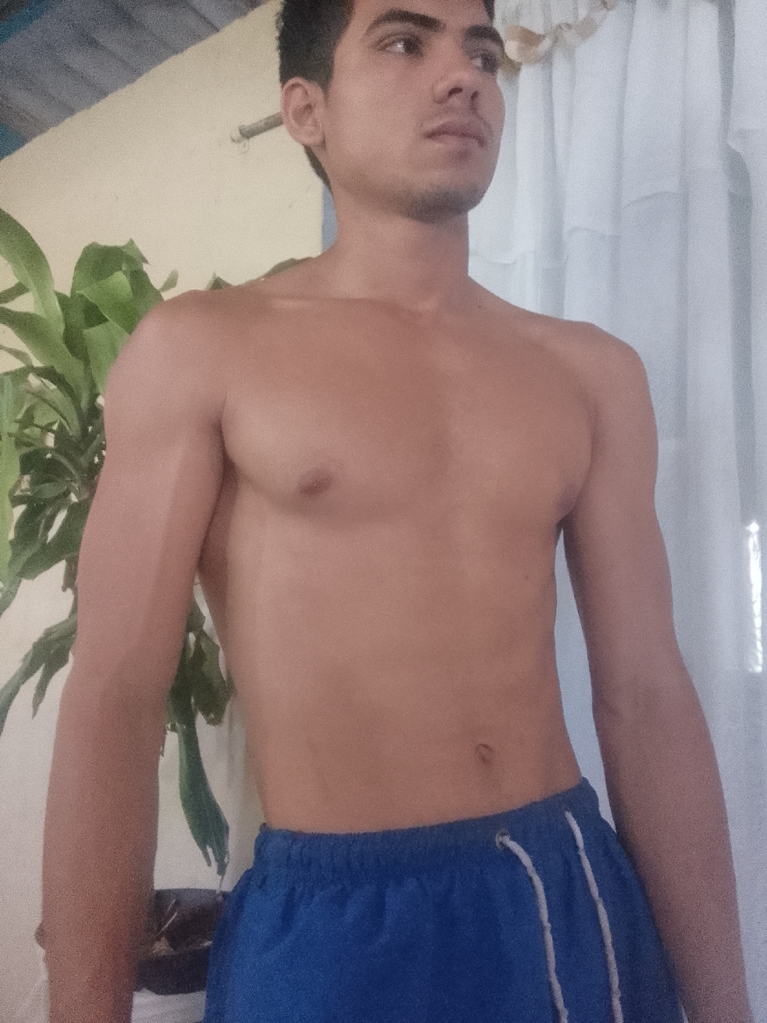 A shirtless young man with short dark hair and a light complexion is standing indoors. He is wearing blue shorts with white drawstrings. Behind him to the left is a green leafy plant, and to the right, a white curtain. A cream-colored wall is visible in the background. The location is Juan Griego, Venezuela.