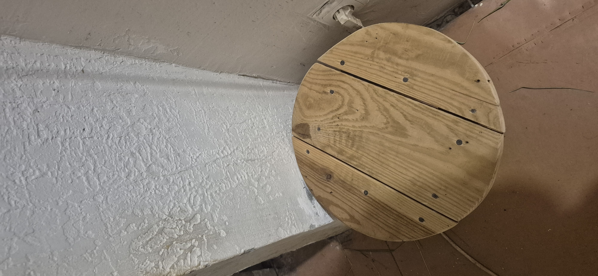 The image shows a circular wooden object with visible wood grain and nail heads, positioned on a brown, tiled floor. Behind the wooden object, there is a rough, textured white wall with some visible cracking and imperfections. To the right of the wall, a cream-colored electrical outlet with a white plug is visible. The scene appears to be indoors, likely in a utilitarian or unfinished space. There is no indication of the city of Kryvyi Rih, Ukraine, or any specific location within it. The image is a still photograph with no observable actions or interactions taking place.