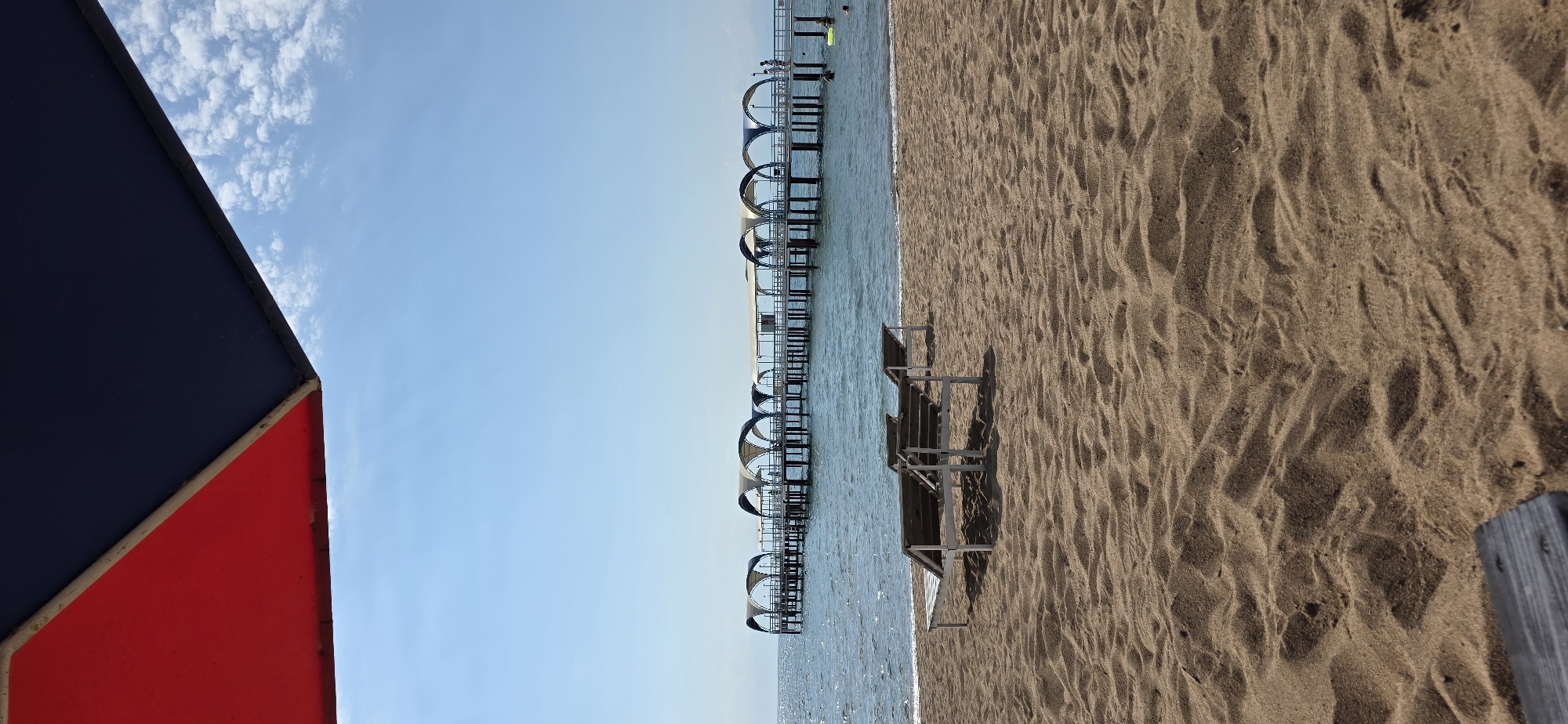 This is a vertical, eye-level shot of a beach in Kara-Oy, Kyrgyzstan, with a clear blue sky and a pier extending into the water. In the foreground, two empty wooden lounge chairs sit on the sandy beach. The sand shows footprints and ripple patterns. A portion of a dark wooden table is visible in the bottom left corner. The middle ground features the calm, blue water of a lake or sea. In the background, a long pier with arched, covered sections stretches across the water. Several people are visible on the pier, some walking and others standing near the water's edge. A few individuals are also in the water, with one person visible on a yellow float. The top of the frame is dominated by a red and dark blue umbrella casting a shadow. The overall scene depicts a tranquil beach setting on a sunny day.