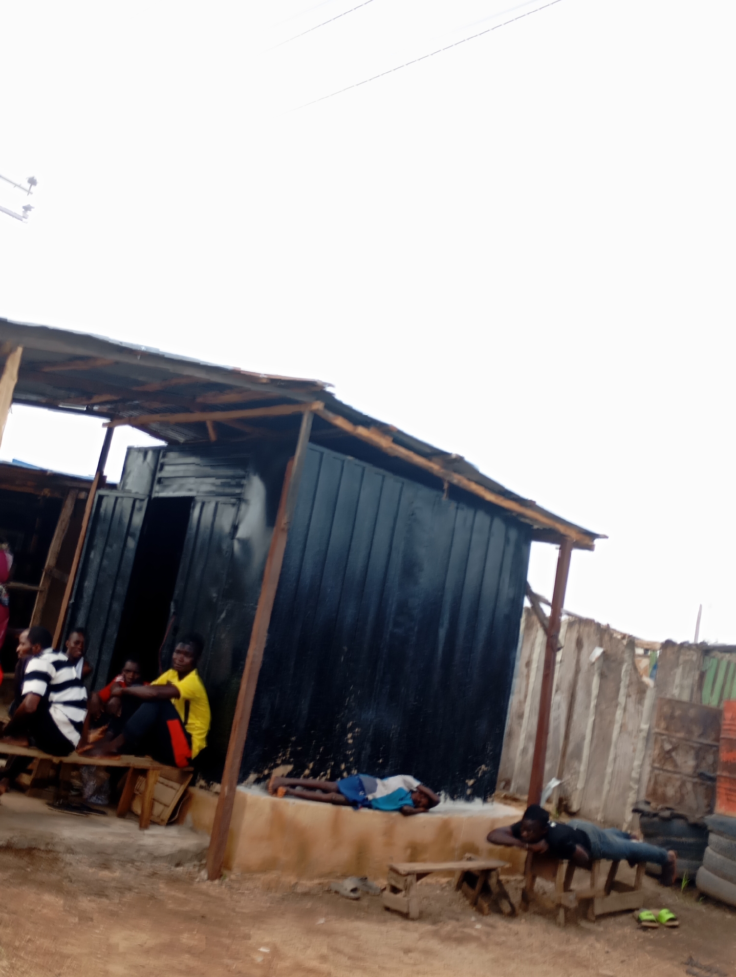 In Shendam, Nigeria, a low-angle, vertical shot captures a scene outside a weathered, corrugated metal structure with a makeshift roof of corrugated tin and wood. In the foreground, a young person lies prone on a wooden bench, head resting on their arms. To the left, a group of four young people sit on another wooden bench. One person wears a black and white striped shirt, another a red shirt, and a third a bright yellow t-shirt with red accents on black pants. A fourth person is partially obscured. The ground is dirt and uneven. Further back, a rough wooden fence and stacked tires are visible. The sky is overcast and bright.