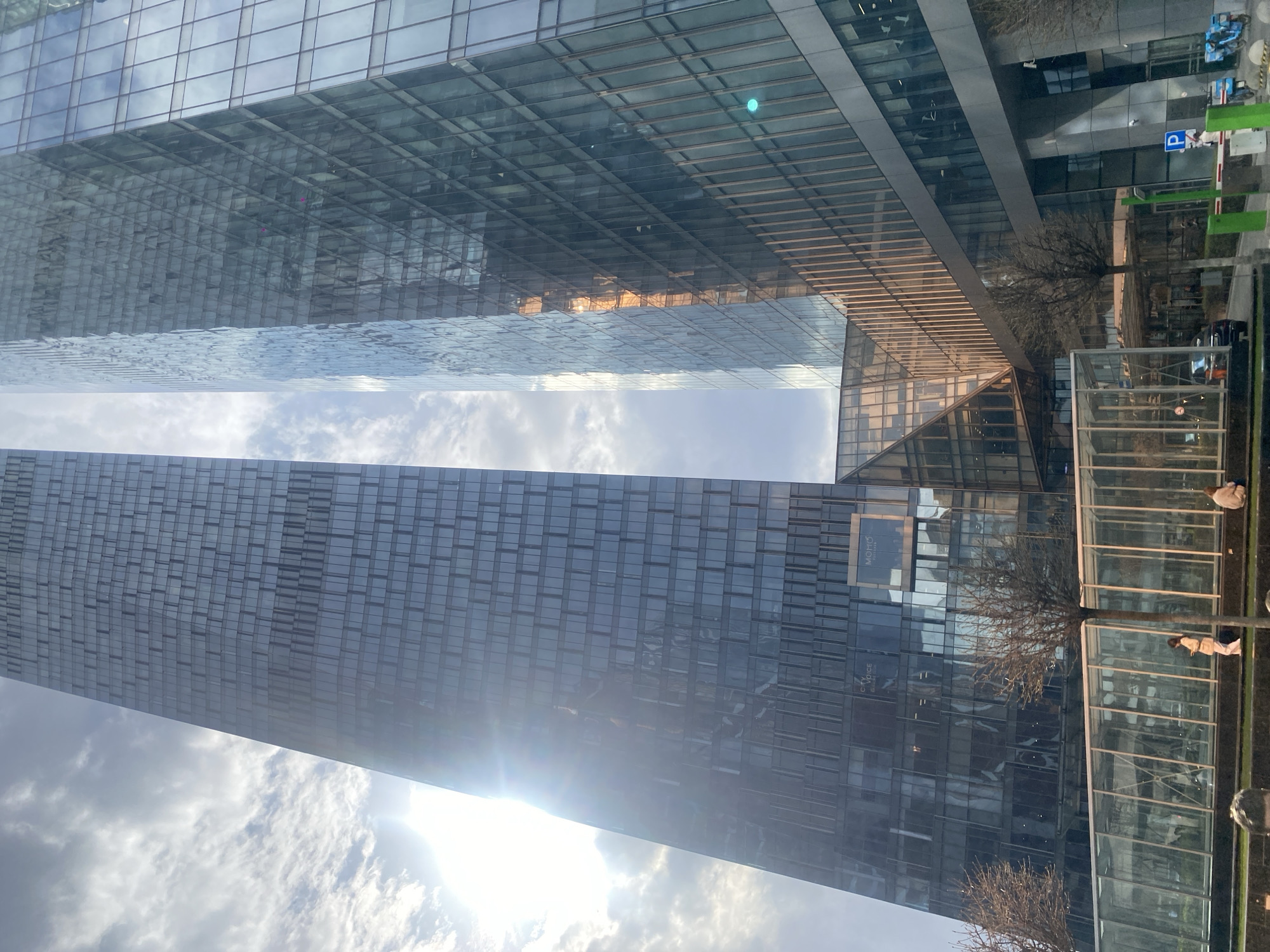 The image shows a cityscape in Moscow, Russia, featuring modern architecture with glass skyscrapers. The view is taken from below, looking up at the buildings, and includes the sky with clouds and sunlight. The buildings reflect the sky and each other. There is a parking barrier with signs. Pedestrians can be seen in the lower right corner, but they are out of focus and not clearly identifiable. Some bare trees are visible.