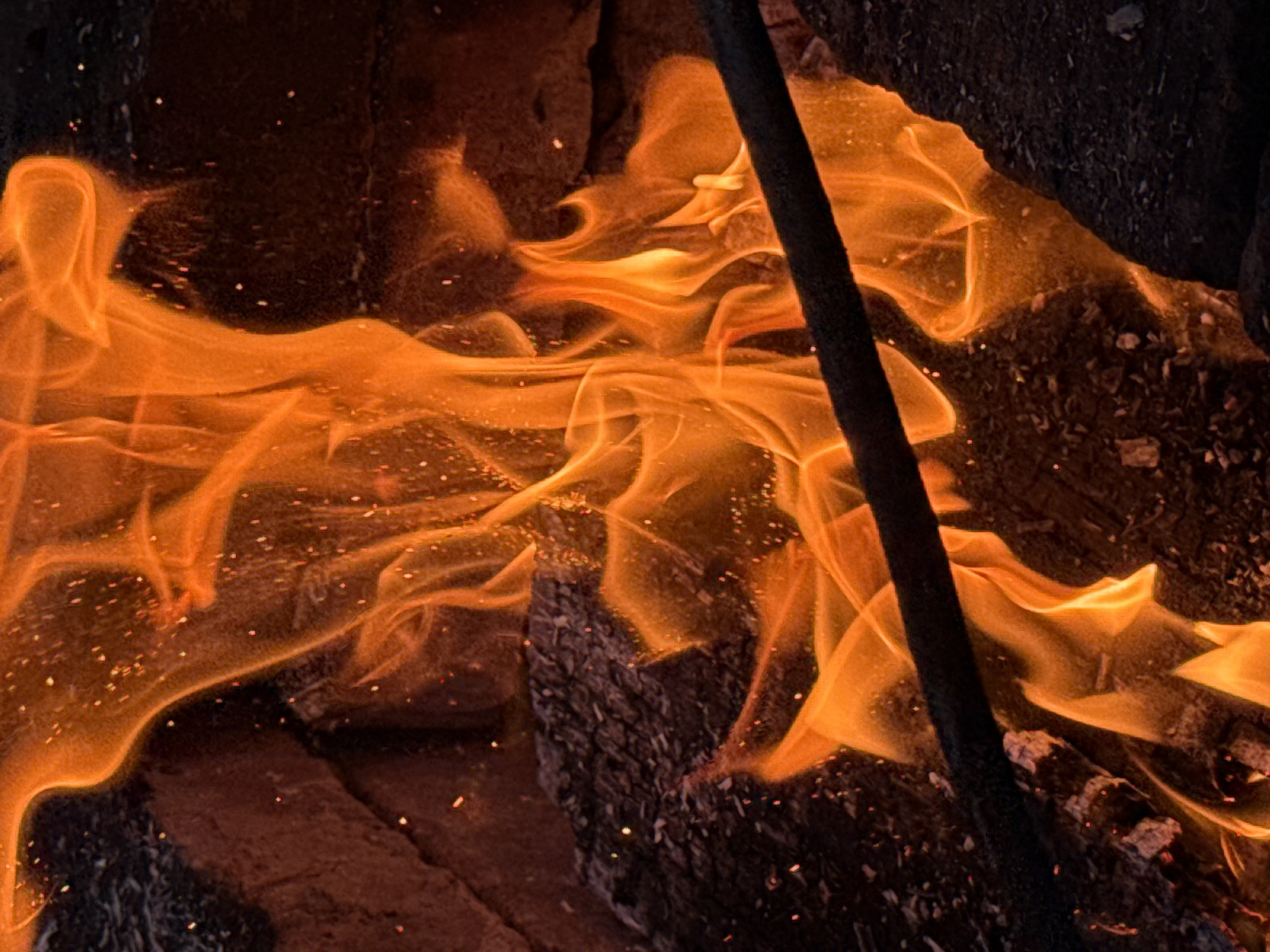 A close-up, dynamic shot captures the intense flames of a fire engulfing logs and bricks. The vibrant orange and yellow hues of the fire are mesmerizing, with sparks scattered throughout, suggesting heat and energy. The rough texture of the logs and the rough, uneven surfaces of the bricks provide a stark contrast to the fluid movement of the flames. A dark, cylindrical object, possibly a metal rod, cuts diagonally across the lower portion of the image, adding a sharp, linear element to the chaotic beauty of the fire. The background is dark and indistinct, emphasizing the focal point of the burning elements. The scene evokes a sense of raw power and primal energy, hinting at a controlled burn or an outdoor fire pit.