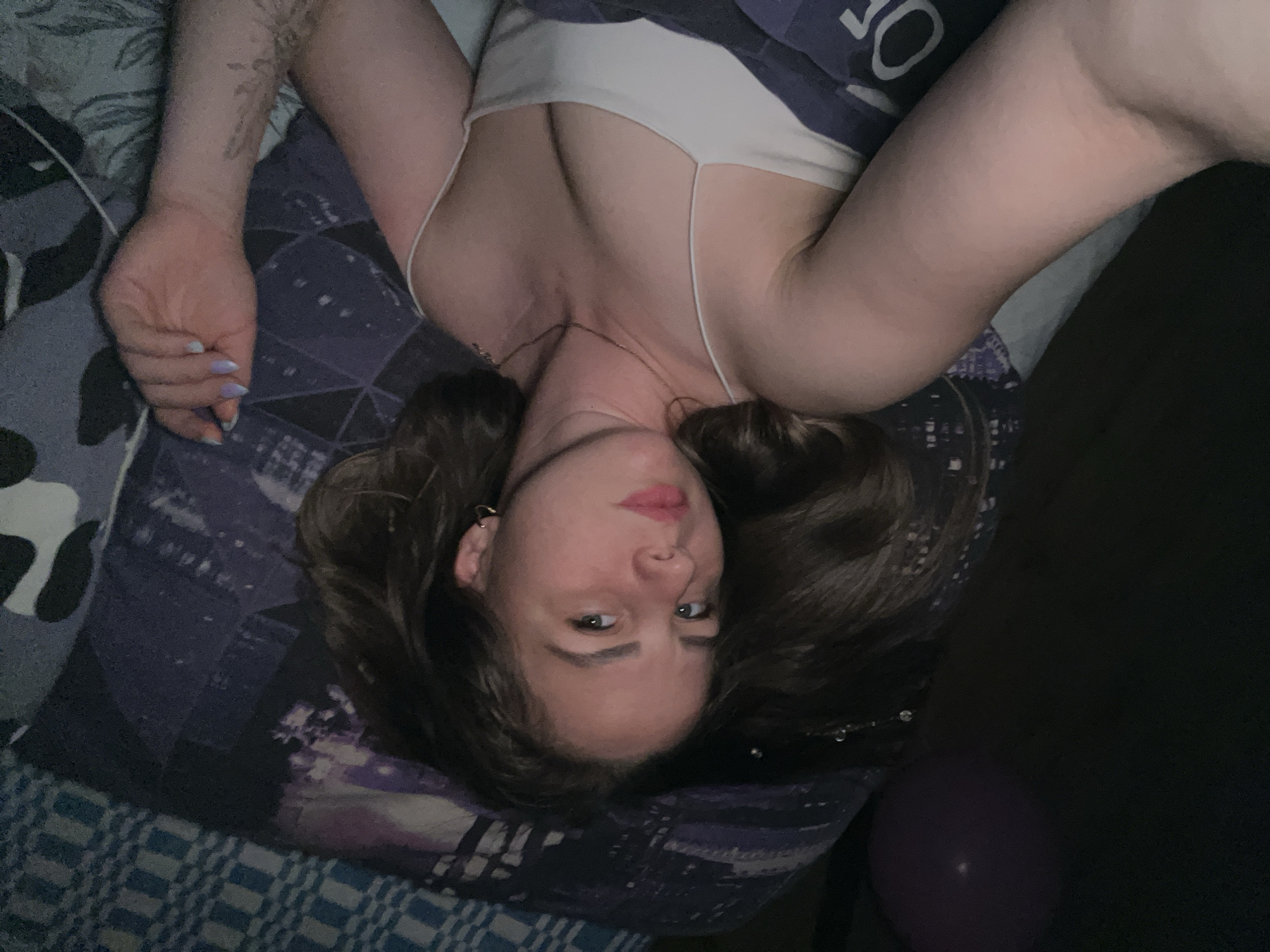 A young woman is captured in a close-up, slightly rotated portrait, lying down and looking directly at the camera. She appears to be on a bed in a dimly lit indoor setting, likely in Izhevsk, Russia.
The woman has long, dark brown hair spread out around her head on a dark blue and purple patterned blanket. She is wearing a light-colored (possibly white or off-white) camisole or tank top with thin spaghetti straps, revealing her décolletage and a delicate necklace. Her facial expression is calm, with light pink lipstick and her eyes focused on the viewer. A small hoop earring is visible in her left ear.
Her arms are extended upwards in the frame, resting on the bedding. On her left forearm, a faded tattoo with a floral or abstract design is visible. Her fingernails are painted with a distinct two-tone design, featuring alternating or gradient light blue/purple and peach/orange shades.
The bedding consists of multiple patterns: a checkered or striped blue and white fabric on the far left, and a dark blue and purple geometric or abstract pattern under and around her. A portion of a dark blue fabric with faint, out-of-focus white text, possibly "JO" or "50", is visible behind her right shoulder. The right side of the image is mostly dark, with a glimpse of a purple balloon in the bottom right corner. The lighting suggests an evening or nighttime shot, with artificial light sources.