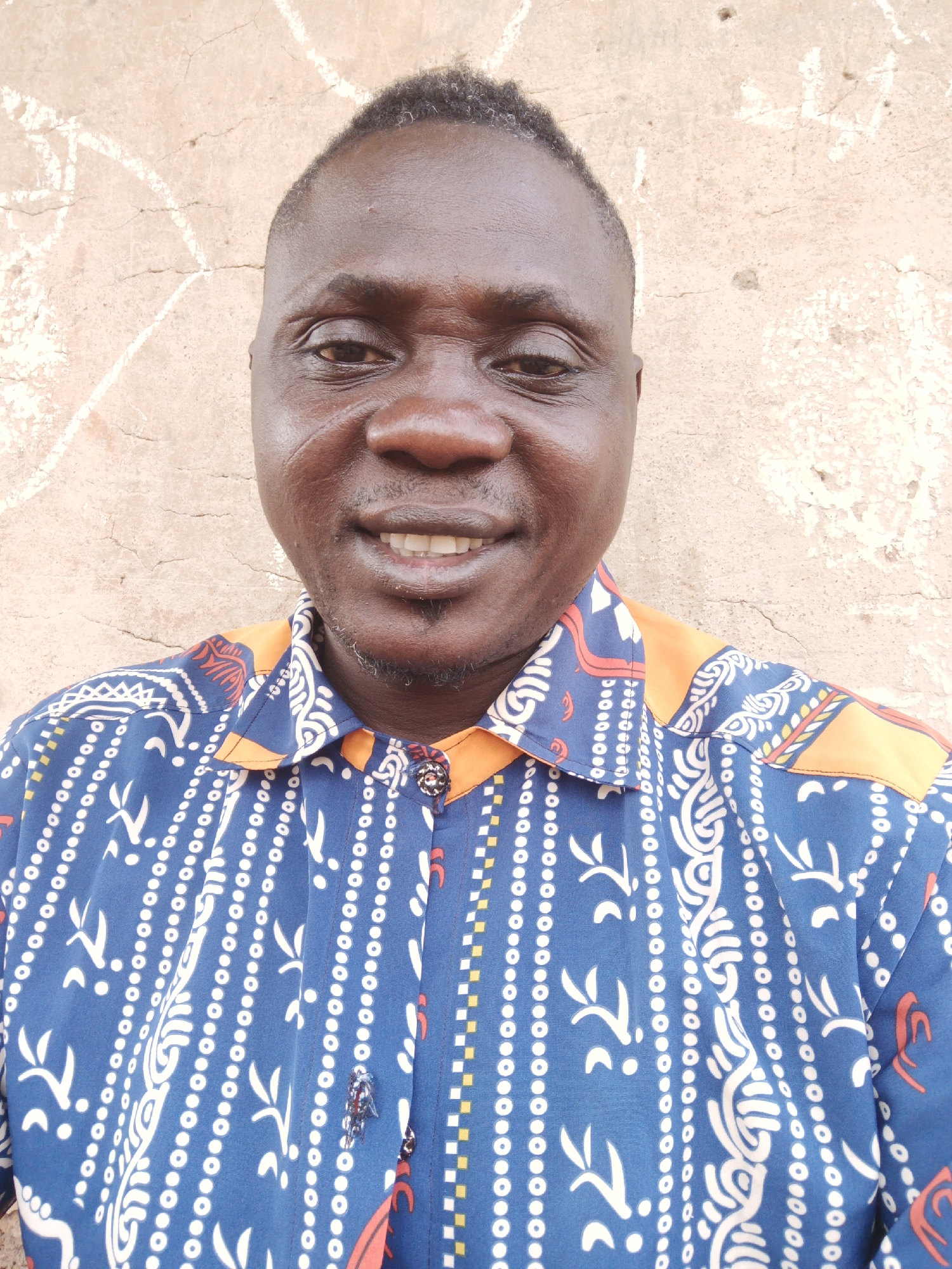 A medium shot depicts a middle-aged Black male smiling at the camera. He has short, dark hair and a goatee. He is wearing a blue patterned shirt with orange accents on the collar and shoulders. The shirt is buttoned. The background is a beige, slightly textured wall, possibly concrete or stucco. Given the prompt, the location is Akwanga, Nigeria.