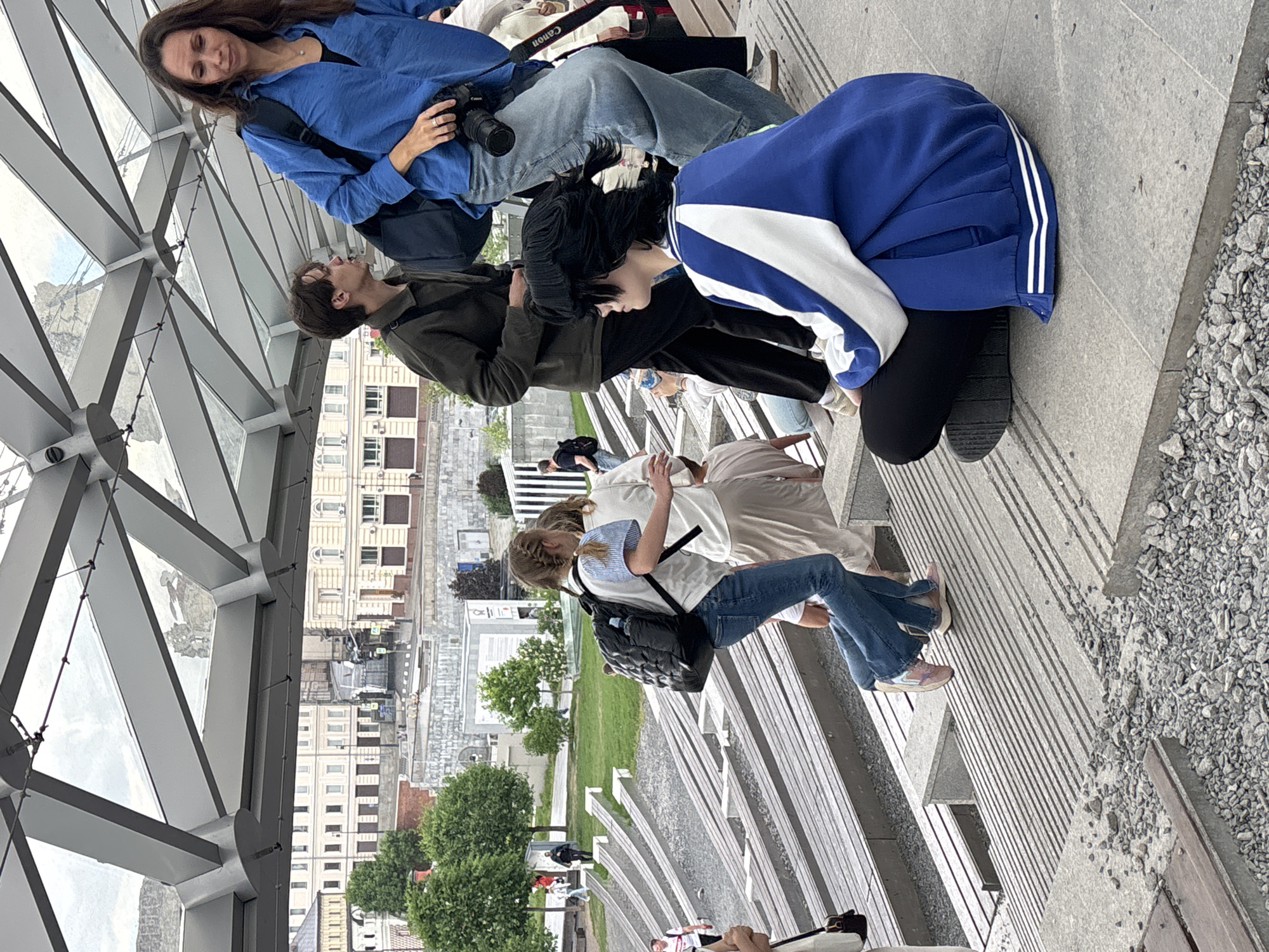 A daytime shot shows a group of people on a structure overlooking part of Moscow, Russia. The foreground features the angled, geometric support structure of the bridge. People are sitting and standing in the center. A woman is wearing a blue button-down shirt and holding a camera. A person in a dark green jacket looks upward. Another person is wearing a blue and white jacket. Below them, another group is walking on stairs. The background shows a cityscape with buildings and green spaces.