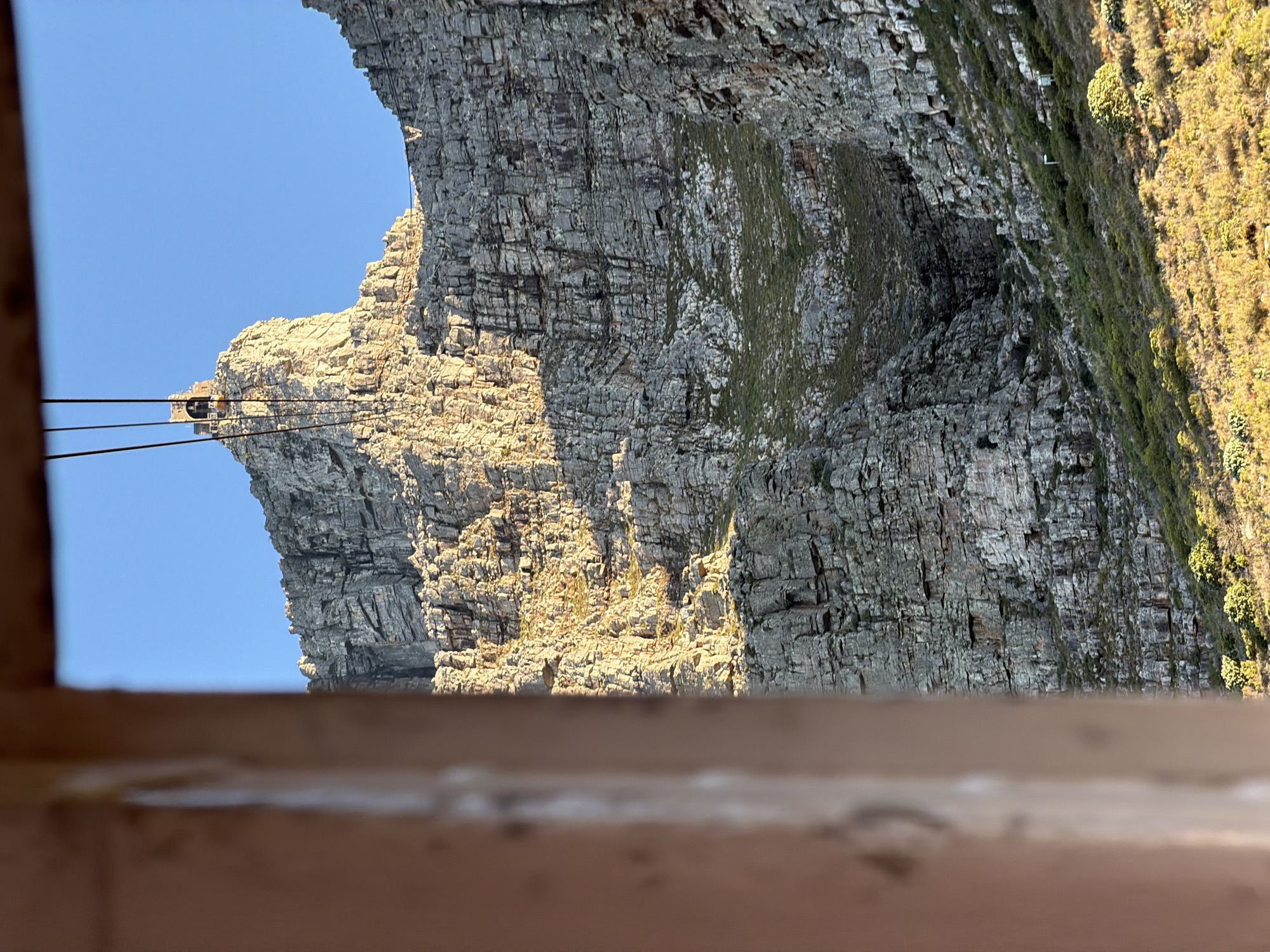 The image displays a dramatic, rugged cliff face under a clear blue sky. The original photograph is oriented on its side; if rotated 90 degrees clockwise, the sky would be at the top and the mountain face would rise vertically.
The scene is dominated by a massive, layered rock cliff, exhibiting deep fissures and varying shades of grey. Patches of green and brown vegetation, including moss and scrub, cling to sections of the rock face and cover a more gently sloping area to the right (if rotated). Golden sunlight brightly illuminates the upper and central portions of the cliff, casting distinct shadows that highlight its rough textures and suggesting either early morning or late afternoon.
On a distant, prominent peak or outcrop towards the left (when correctly oriented), a small building or station, likely part of a cable car system, is visible. Multiple dark cables extend horizontally from this structure across the clear blue sky towards the viewer's implied position. The viewpoint appears to be from within an enclosed space or on a platform, as a blurred, light-brown wooden or metallic railing occupies the bottom foreground of the image.
No people or discernible text are visible. The majestic natural landscape, combined with the cable car infrastructure and layered geological formations, strongly indicates a location such as Table Mountain in Cape Town, South Africa. The weather is clear and sunny.