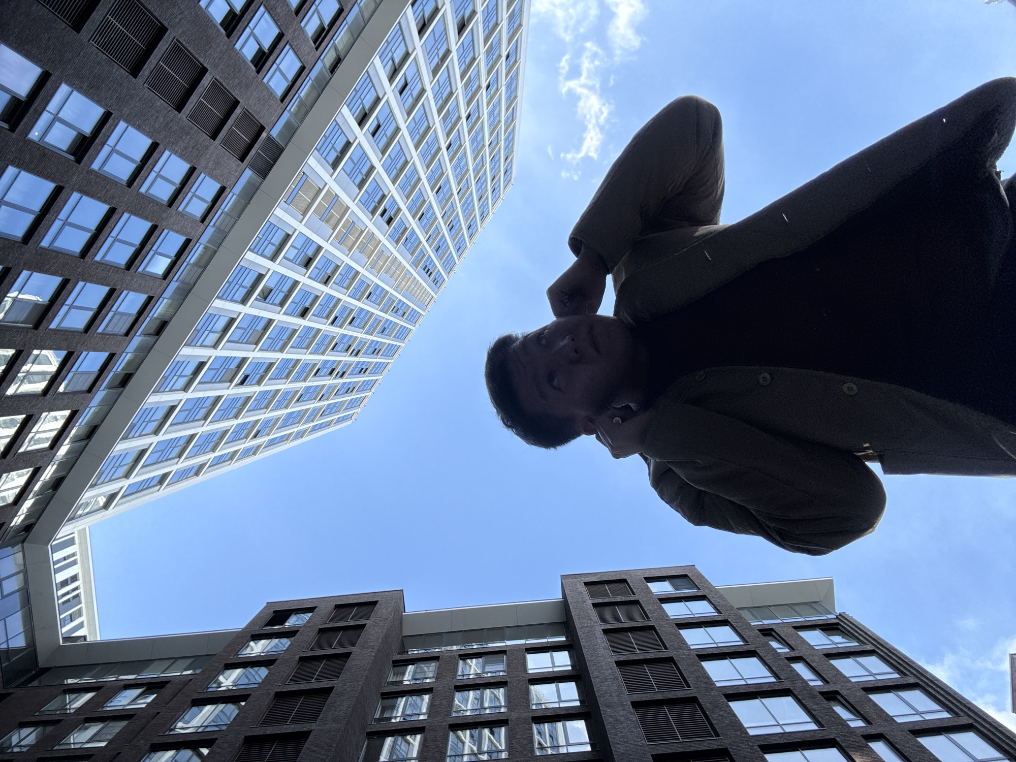 The image depicts a person looking up at tall buildings against a blue sky. The buildings are modern, with many windows, reflecting the sky. The person is positioned in the lower part of the frame. The photograph was taken in Moscow, Russia.