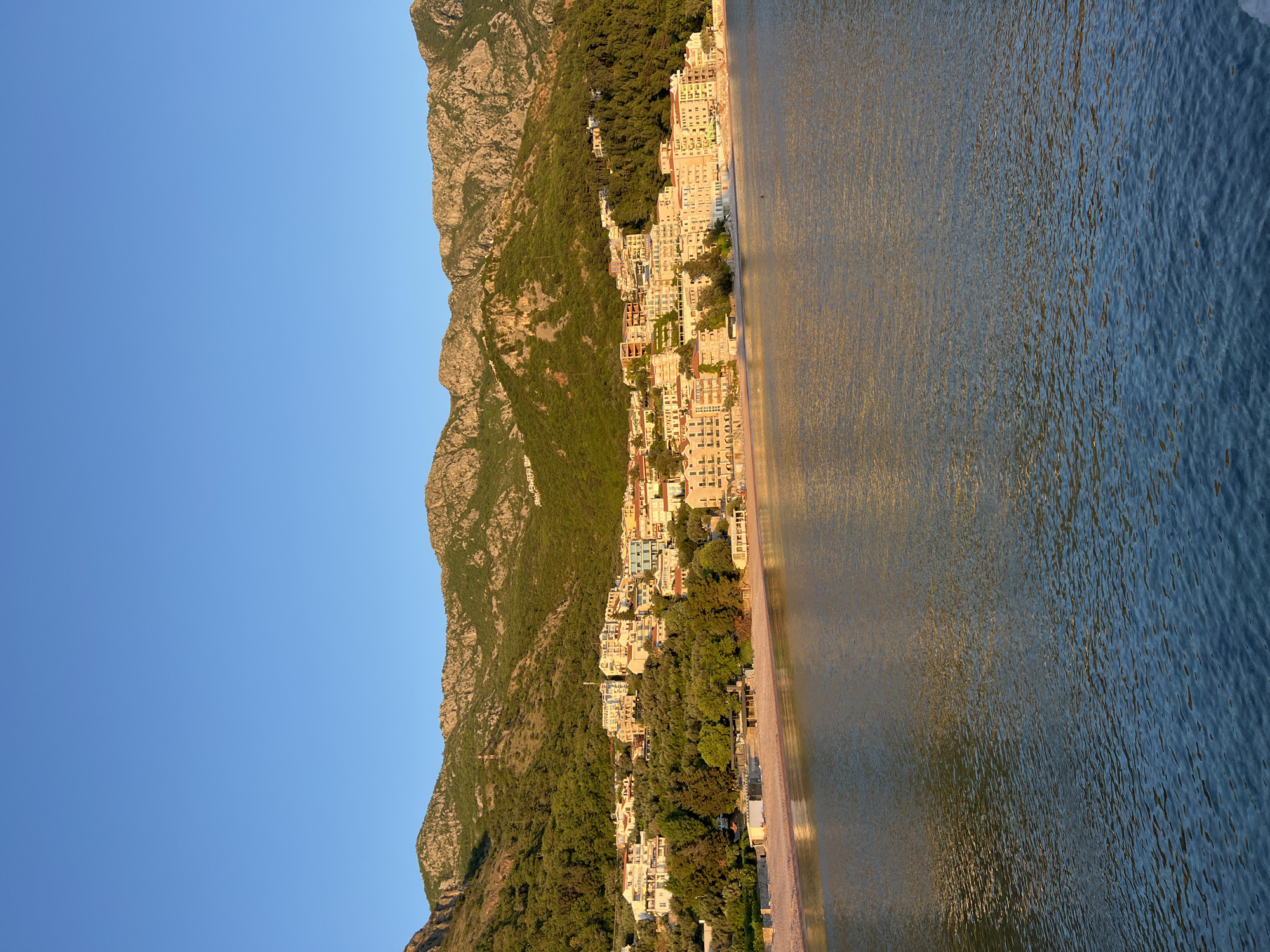 A panoramic view captures a picturesque coastal scene, likely Sveti Stefan, Montenegro, at either sunrise or sunset, evidenced by the warm, golden light bathing the landscape. The scene is dominated by a majestic, steep mountain range on the left, covered in a mix of dense green foliage and rugged, rocky outcrops. Along the base of this mountain, a charming town stretches along a narrow coastline, featuring numerous light-colored buildings – mostly multi-story structures with balconies and flat roofs, possibly hotels or residences.
To the right of the town and beach, a calm sea shimmers under the low sun, its surface reflecting a gradient of deep golden-orange closer to the shore, transitioning into a darker blue further out. Subtle ripples are visible on the water. A narrow strip of beach separates the buildings from the sea. The sky above is a clear, cloudless blue, indicating fair weather. No people or discernible activities are visible, and there is no visible text. The overall impression is one of serene natural beauty combined with a well-integrated coastal settlement.