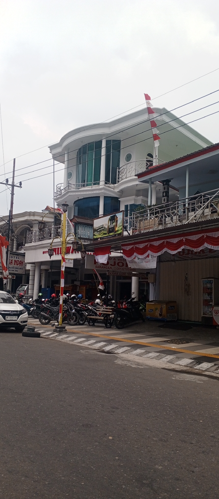 The image captures a street scene in Sarangan, Indonesia, featuring a multi-story building with a curved facade, balconies, and large windows, topped with an Indonesian flag. In front of the building, a row of parked motorcycles is visible, suggesting a commercial area. Banners and signs adorn the building's lower levels, indicating businesses such as a hotel and possibly other services. A white car is parked to the left of the frame. The street is paved, with a pedestrian walkway delineated by yellow and black markings. The sky is overcast.