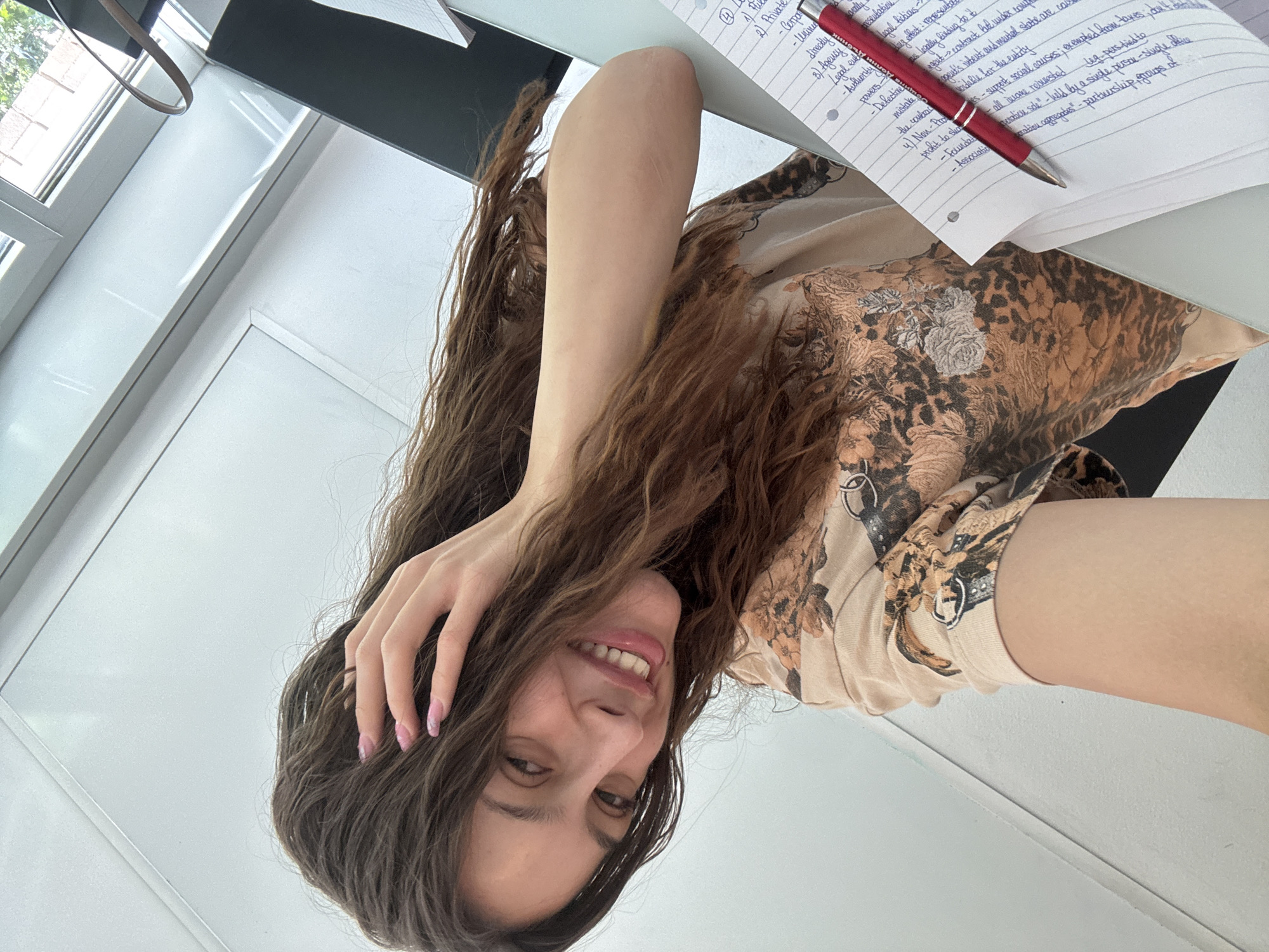 A young woman with long, wavy brown hair is smiling at the camera while sitting at a desk. Her right hand is in her hair. On the desk is a notebook filled with handwritten notes and a red pen. The notes appear to be legal or business-related. The background includes a window. Given the writing on the page, the location is likely a home office or study area, as it is unlikely someone would be studying in a public place in Milan, Italy.