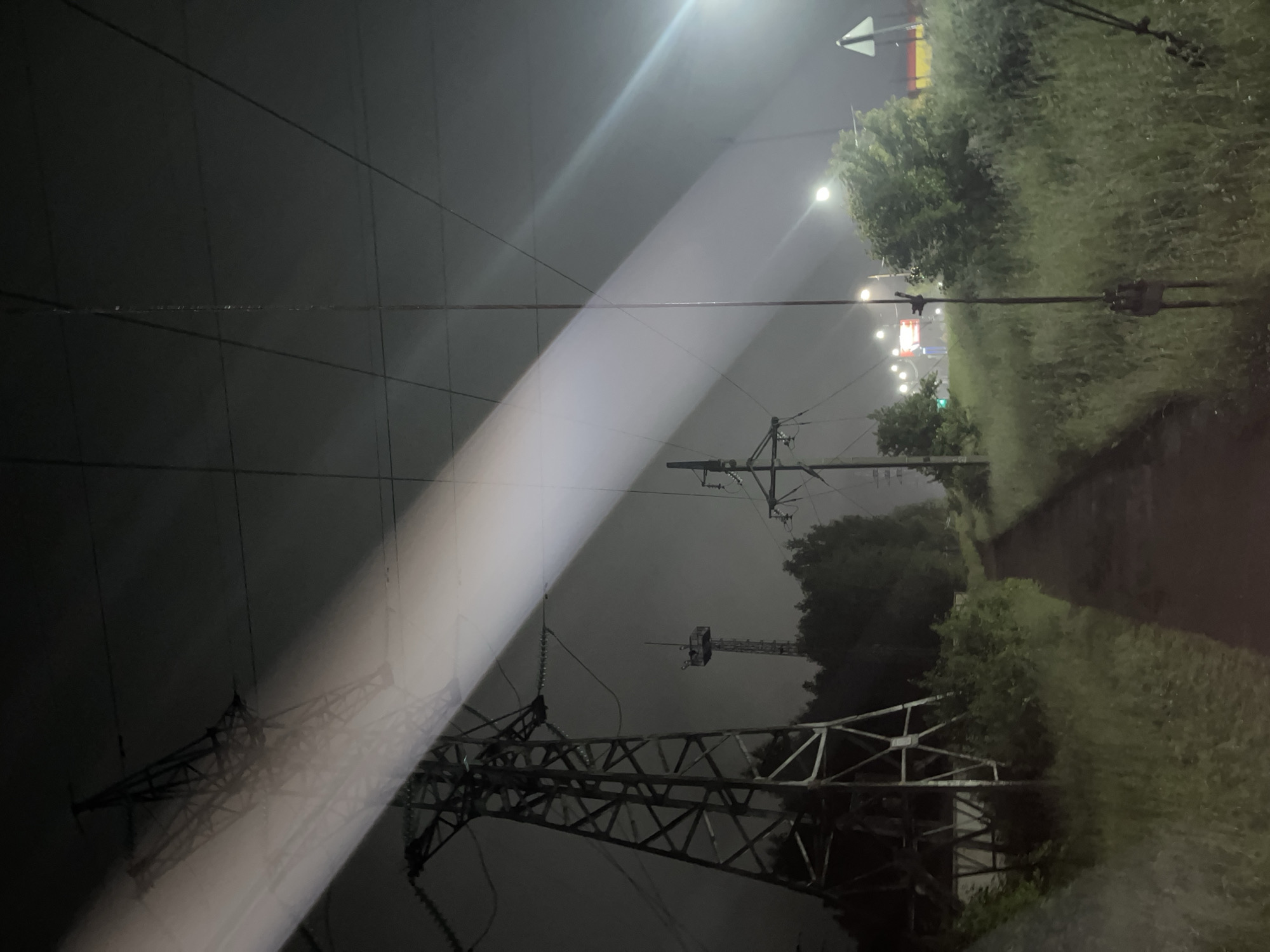This nighttime image, captured in Chaikovsky, Russia, presents an outdoor scene characterized by industrial infrastructure and prominent atmospheric lighting. A broad, intense white light beam traverses the upper half of the frame diagonally, originating from the upper left corner and extending towards the lower right. This beam strongly illuminates the atmosphere, suggesting the presence of mist, fog, or low clouds.
In the left midground, a substantial metal lattice-work high-voltage transmission tower is visible, its structure partially overlaid by the bright light beam. Multiple insulators and power lines are discernible extending from the tower. Further to the right and positioned deeper in the scene, another, smaller power pole is partially illuminated by the diagonal light. Overhead, numerous thin, dark power lines crisscross the upper portion of the sky. A slender, dark vertical pole or antenna extends from the bottom center-right of the frame upwards, nearly reaching the top edge.
At ground level, an unpaved path or dirt track runs from the lower center towards the mid-right background. Lush green grass and dense bushes line both sides of this path and extend into the foreground. A contiguous line of dark trees forms a backdrop behind the power infrastructure and along the horizon line.
In the left background, behind the large power tower and obscured by trees, a tall, slender lattice-work communications or observation tower with an elevated platform is visible. In the mid-right background, several bright, circular streetlights or artificial light sources are present, casting distinct white and yellowish glows. Faint red and blue lights are also discernible in the far distance. A white triangular sign is partially visible on the far right side of the frame, situated within the foreground vegetation. No individuals are present within the scene.