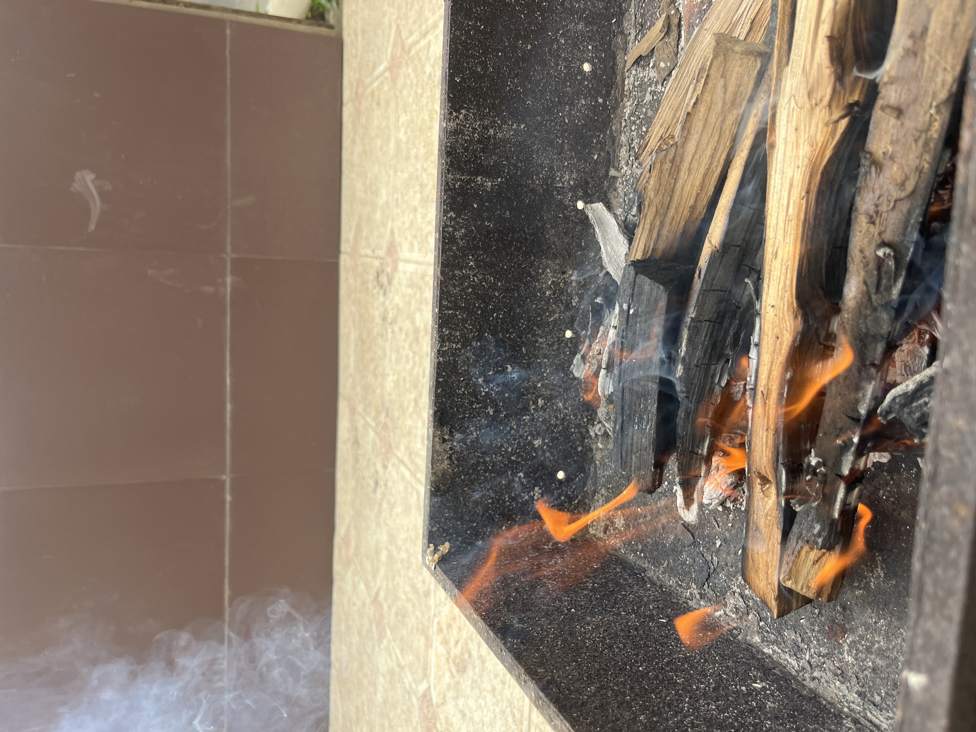 The image shows a close-up view of a barbecue grill filled with burning wood and embers. Wisps of smoke rise from the grill, obscuring a tiled wall in the background and a tiled floor in the foreground. The grill appears to be located outdoors, possibly in a residential or public area. There are no people visible in the image. The focus is on the fire, emphasizing its heat and texture. The scene is set in Kaspiysk, Russia, suggesting a potentially cold climate where outdoor fires might be used for warmth or cooking.