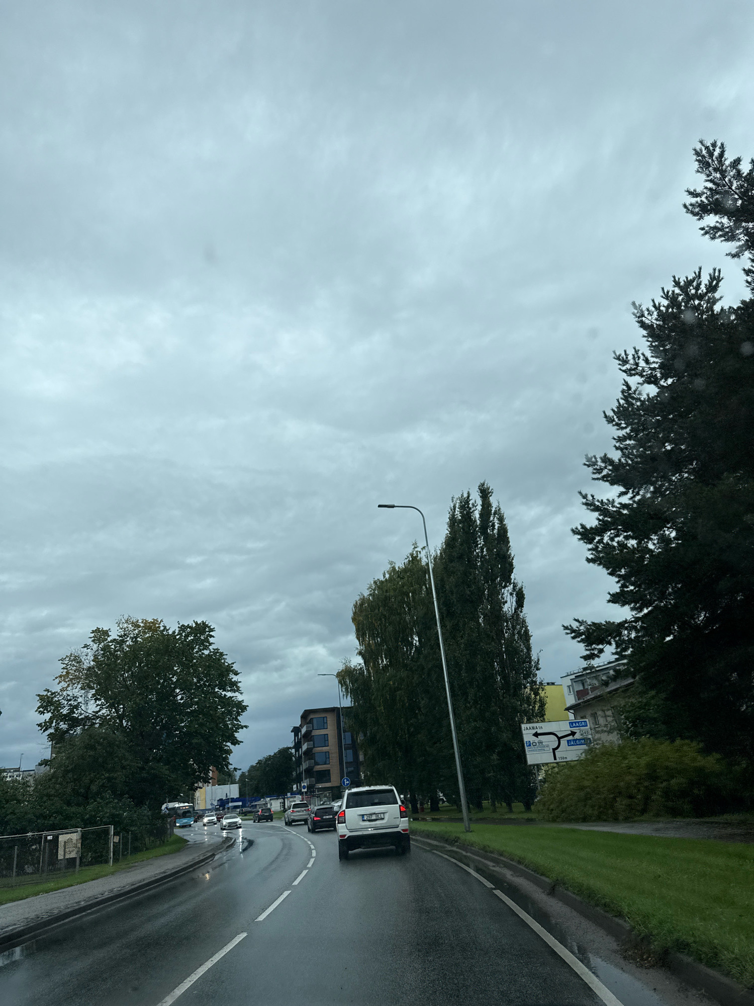 The image captures a view of a wet asphalt road in Saku, Estonia, on a cloudy day. Multiple vehicles are present on the road, including a white SUV in the foreground, a white sedan further ahead, and other cars in traffic. Trees line the sides of the road, with some appearing tall and slender, while others are fuller. Buildings are visible in the background, including apartment buildings and a smaller structure. A street sign with directional arrows indicating "JÄÄMA tn" and "LAAGRI" is positioned on the right side of the road, suggesting a junction or intersection. The ground appears wet, indicating recent rainfall, and puddles are visible on the road. The sky is overcast with a significant cloud cover.