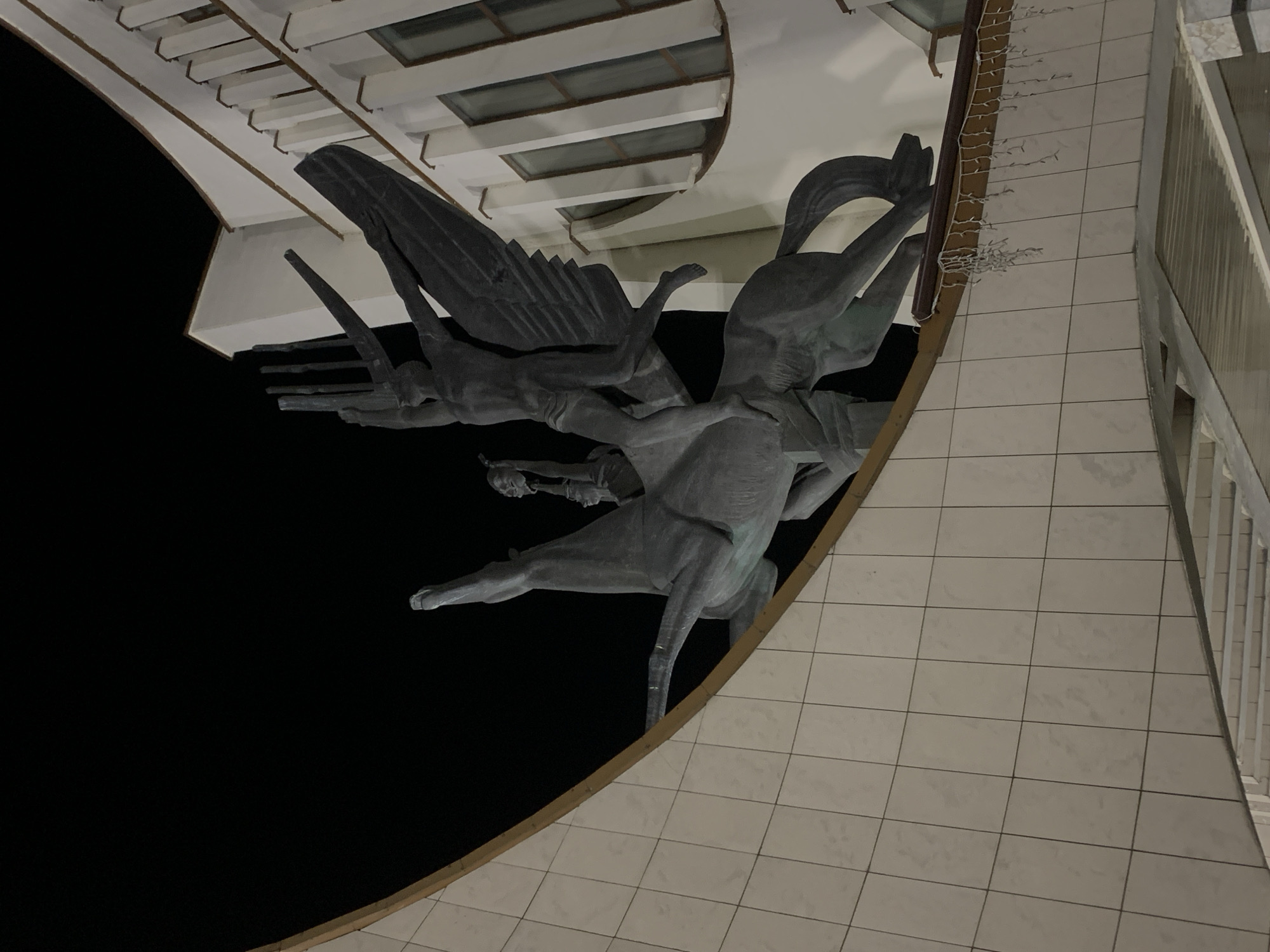 The image shows a sculpture affixed to the side of a building, likely in the city of Hrodna, Belarus. The sculpture depicts a scene with multiple figures, including winged entities and at least one horse. The figures are rendered in a dark, metallic tone. The sculpture is mounted on a building with a light-colored, tiled facade and a portion of a railing is visible. The backdrop to the left of the sculpture is a solid black, suggesting nighttime or a dark interior space. The building has white trim and architectural details.