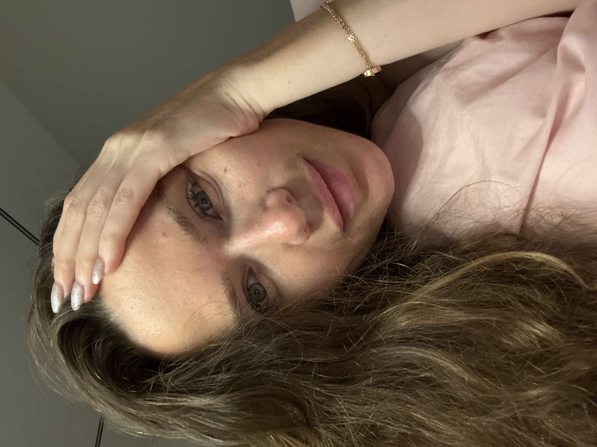 In a selfie taken in Khimki, Russia, a woman with long brown hair rests her forehead on her hand, her eyes looking at the camera. She is wearing a pink top and a gold bracelet on her wrist. The background is plain and indistinct.