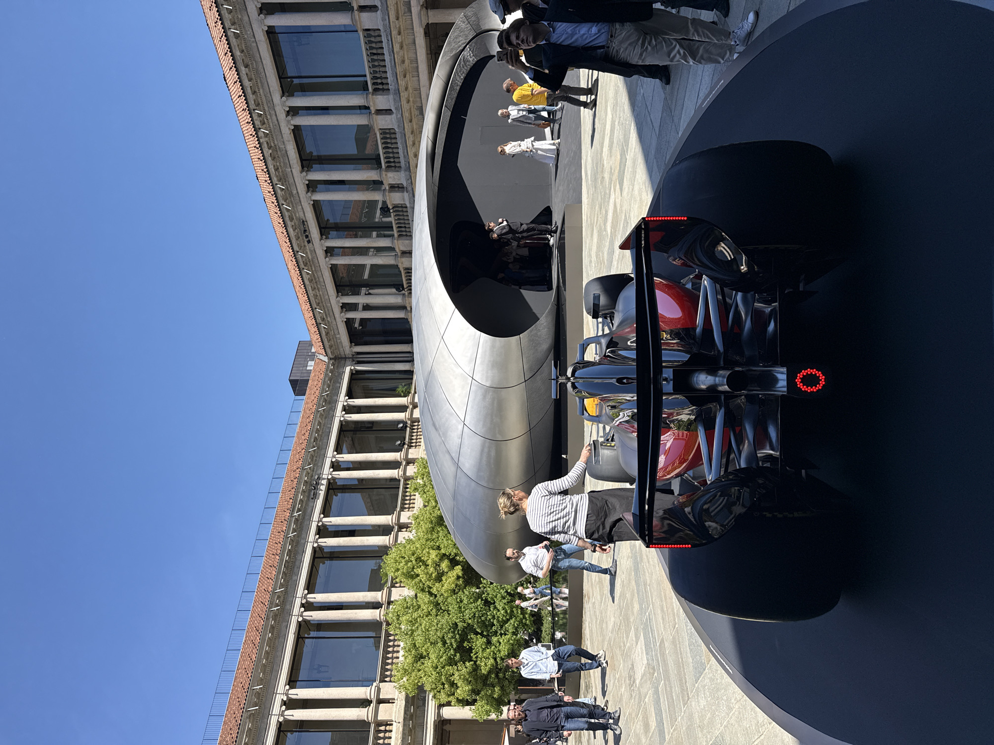 The image captures an outdoor scene in what appears to be a public square or courtyard, under a bright, clear blue sky. A prominent feature is a sleek, modern, silvery-grey tubular structure with a large elliptical opening, possibly an exhibition pavilion or art installation.
In the foreground, positioned on a dark, circular platform, is a high-performance racing car, likely a Formula 1 vehicle. It is predominantly black with red accents, viewed from the rear, showcasing its intricate aerodynamic components, a large rear wing with integrated red LED lights, and a circular red brake light.
To the left of the modern structure stands a multi-story building featuring classical architecture, characterized by rows of columns, arches, large windows, and reddish-brown brickwork with lighter stone details. A small tree with vibrant green foliage is visible between the building and the modern structure.
Numerous people are present in the scene, observing the car and the contemporary structure, or simply walking around. Their attire suggests a casual public setting. The bright daylight indicates a sunny day, consistent with an outdoor event. No discernible text is visible on any of the objects or structures. Given the context, this scene is set in Milan, Italy, blending historical architecture with modern design and an automotive exhibition.