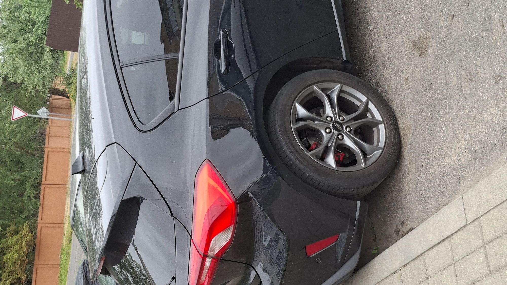 The image shows a black Ford Focus parked on a street in the city of Smolevichi, Belarus. The car is viewed from the rear quarter, with the rear passenger-side wheel and tail light clearly visible. The car is dark and appears to be in good condition, with a shiny finish. The wheel is a dark gray alloy with red brake calipers. The tail light is a sharp, angular design with red and clear sections.
The car is parked on the right side of the street, next to a sidewalk made of light-colored pavers. The street itself is asphalt, with some stains and debris visible. In the background, there is a low, reddish-brown fence, followed by trees and foliage. A street sign with a white triangle and red border is visible in the distance. The environment appears to be a residential or suburban area. There are no people visible in the image. The lighting suggests it is daytime, likely overcast or late afternoon, as there are no harsh shadows.