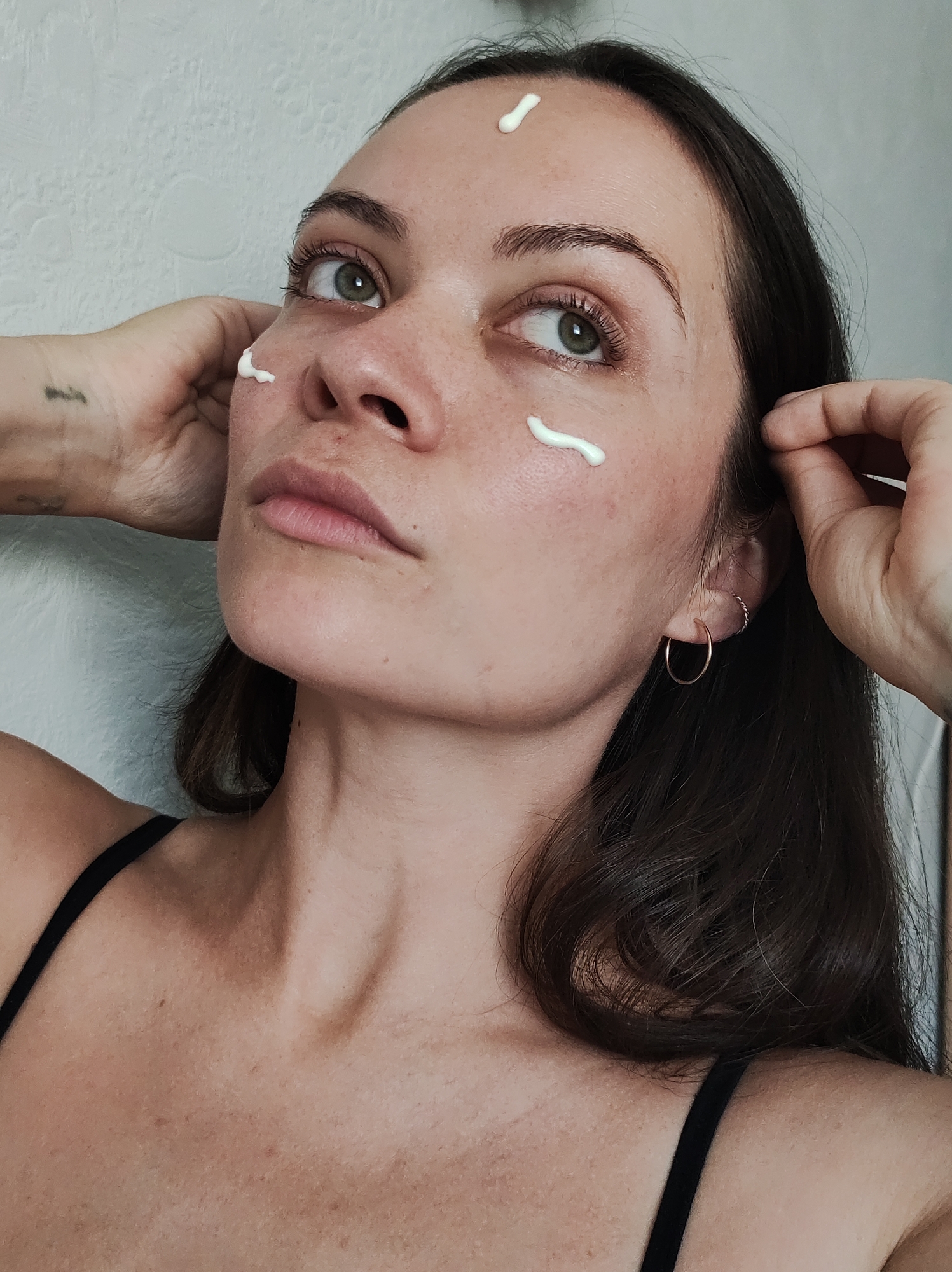 A woman with fair skin and dark hair is shown in a medium shot, looking upwards and to the right. She has three visible dabs of a white, opaque cream-like substance on her face: one on her forehead, and one on each cheek near her eyes. Her hands are raised towards her head, with her fingers lightly touching her hair. She is wearing a black tank top and small gold hoop earrings. A tattoo is visible on her left hand. The background is a neutral, light-colored wall with a subtle texture. The setting appears to be indoors. The location, Chaikovsky, Russia, is not evident in the image.