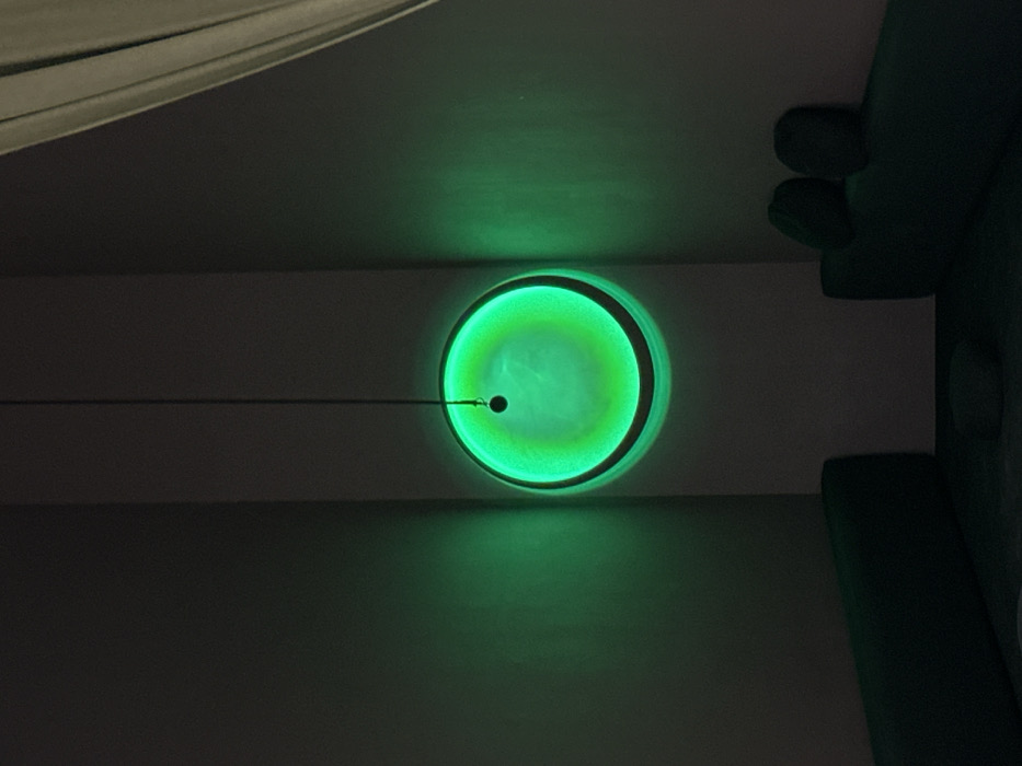 A green circular light fixture is mounted on a flat, dark-colored surface. A thin cord extends horizontally from a small, dark ball hanging in the center of the light. A portion of the ceiling is visible at the top of the image, and an object with rounded edges is visible on the right side. The light emits a bright, somewhat diffused green glow, creating a distinct halo effect on the surrounding surface.