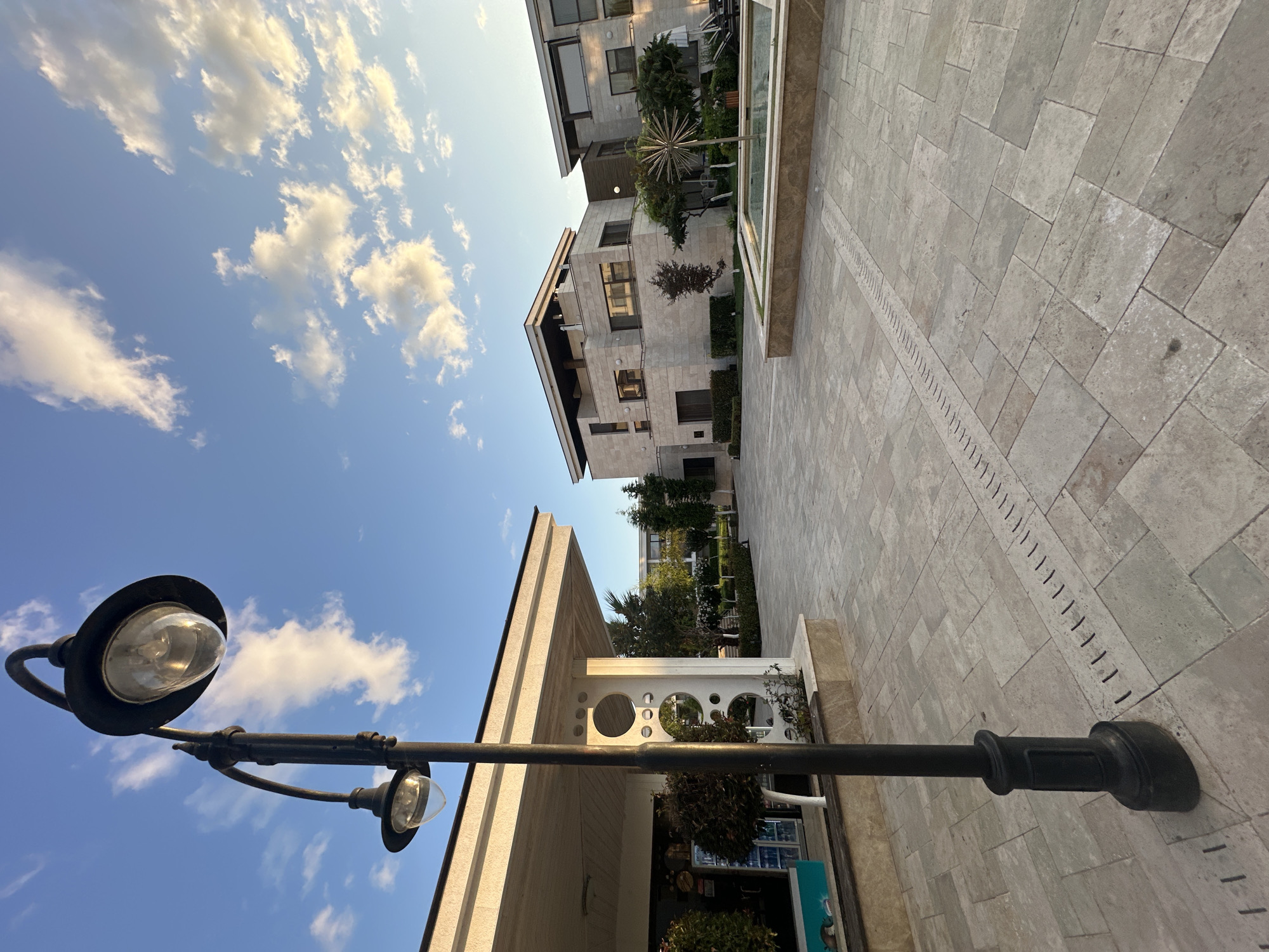 An exterior low-angle shot captures a modern courtyard with a dual-headed lamppost in the foreground. The lamppost is black, with two glass lamps, one above the other. Behind the lamppost, a pale stone building with a flat roof and a covered walkway is visible on the left. To the right, a contemporary multi-story building made of pale stone with dark-framed windows stands under a bright blue sky with scattered white clouds. In the middle ground, a rectangular fountain with a decorative metallic sculpture is present, surrounded by greenery and paved pathways. The courtyard pavement consists of large, light-colored stone tiles, with a linear drain running through the center. The scene appears to be in the city of Sozopol, Bulgaria.