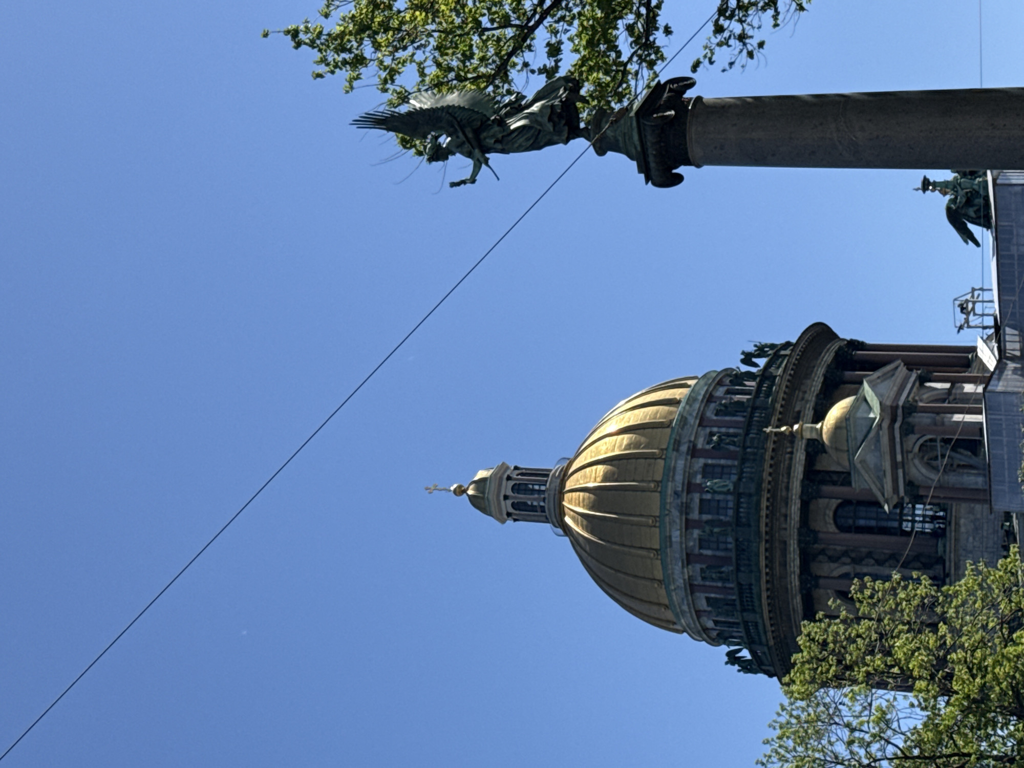 The image depicts a view of the Saint Petersburg cityscape, capturing St. Isaac's Cathedral with its golden dome. In the upper portion of the image is the Alexander Column adorned with a statue of an angel at the top. Some tree branches are visible among the buildings. A diagonal power line traverses the blue sky.