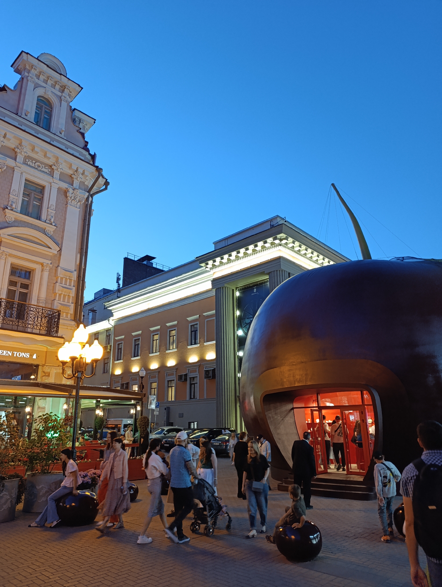 This image captures a lively urban pedestrian scene at dusk or early evening, characterized by artificial lighting contrasting with a deep blue sky. The location is identified as Stoleshnikov Pereulok (Stoleshnikov Lane) in Moscow, Russia, primarily due to the distinctive large, dark, spherical building, commonly known as the "Cherry" building, which is prominently featured on the right side of the frame.
The central subject on the right is the unique, oversized dark brown sphere, resembling a cherry, complete with a long, slender, light-colored stem extending upwards into the sky. This structure functions as a building, with a recessed entrance area displaying a vibrant red interior illumination and glass doors through which several individuals are visible.
To the left, a multi-story building constructed in a classical architectural style features ornate white detailing, including cornices, pilasters, and decorative window surrounds. A balcony with an intricate black iron railing is visible on the second floor. Partial lettering "EEN TONS" is discernible on a sign below this balcony, suggesting a commercial establishment.
In the mid-ground, a warm-toned building with multiple regularly spaced windows and illuminated facade lighting stands behind the cherry structure. Further back, another classical-style building with columns and a lighted roofline is visible. Rooftops with various structures and antennae are present against the sky.
The scene is populated by numerous individuals of varying ages, predominantly adults. Many are casually dressed and are seen walking across the paved pedestrian area. One woman is seated on a smaller, dark spherical sculpture resembling the main cherry building, located on the far left. Another child is sitting on a similar smaller sphere on the right side of the entrance to the cherry building. People are observed entering and exiting the cherry building, while others walk past or engage in social interaction. A street lamp with multiple glowing globes emits warm light on the left, illuminating part of the pedestrian path. Parked vehicles are visible in the background to the right. The ground is paved with interlocking tiles.