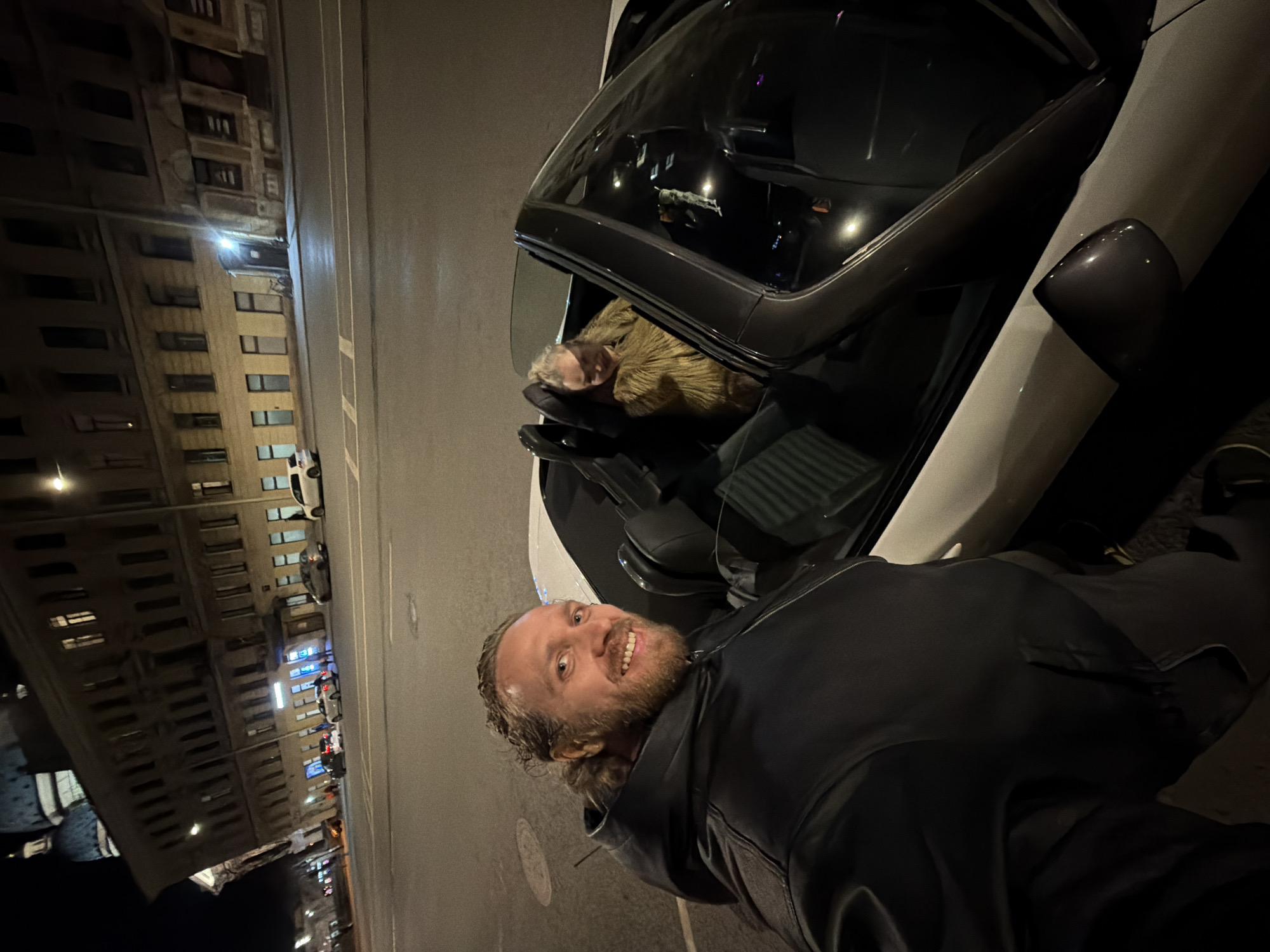 At night, on a street in Saint Petersburg, Russia, a man is posing for a photo in front of a white car with its driver's side door open. A woman is reclining in the driver's seat. Buildings line the opposite side of the street, illuminated by streetlights. Cars are parked along the road. The man is wearing a black jacket and smiling at the camera. The woman inside the vehicle has light hair and is wearing a brown jacket.
