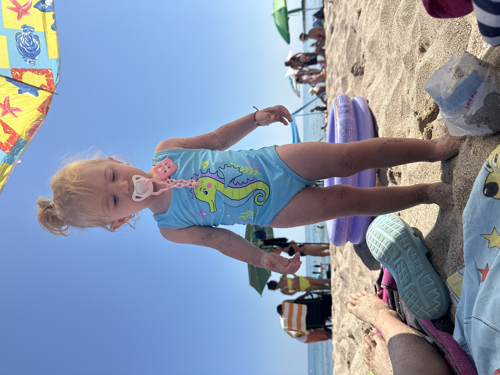 This is a vertical, eye-level, medium shot of a young child on a sandy beach. The child is wearing a light blue swimsuit with a seahorse design and a pink pacifier attached by a pink chain. The child's face is turned to the side and they appear to be looking down, with a somewhat sleepy or contented expression. Their skin is lightly tanned and dusted with sand.
In the background, other beachgoers are visible, some sitting under umbrellas or lying on towels. The sea can be seen in the distance under a clear blue sky. The lighting suggests it is daytime, likely sunny.
Objects visible include a purple inflatable ring near the child, a blue sandal lying on the sand, and a small white plastic bag with some text on it. The text on the bag appears to be "hinten" in blue lettering, which is German for "behind". A colorful beach umbrella is partially visible in the upper left corner of the frame. The overall scene depicts a relaxed day at the beach.