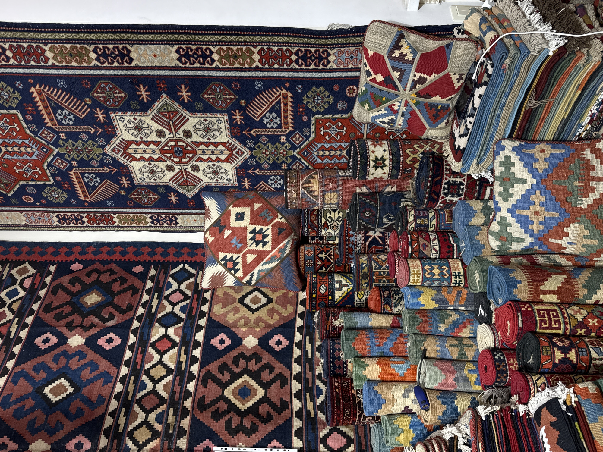 The image shows a collection of rugs and cushions with intricate geometric patterns. A large rug with a dark blue background and geometric designs including star shapes and angular borders occupies the top-left area. Below it is another rug with alternating bands of geometric motifs in shades of dark blue, brown, black, and pink. Stacks of rolled rugs and cushions are piled to the right, showcasing a variety of patterns and colors. The environment suggests a retail setting, possibly a shop or market specializing in handmade rugs and textiles. Based on the location information provided, it is likely this scene is within the city of Yerevan, Armenia, where traditional rug making is a prominent craft. There are no people visible in the scene, and the focus is entirely on the merchandise. The style is traditional Armenian textiles and carpets.