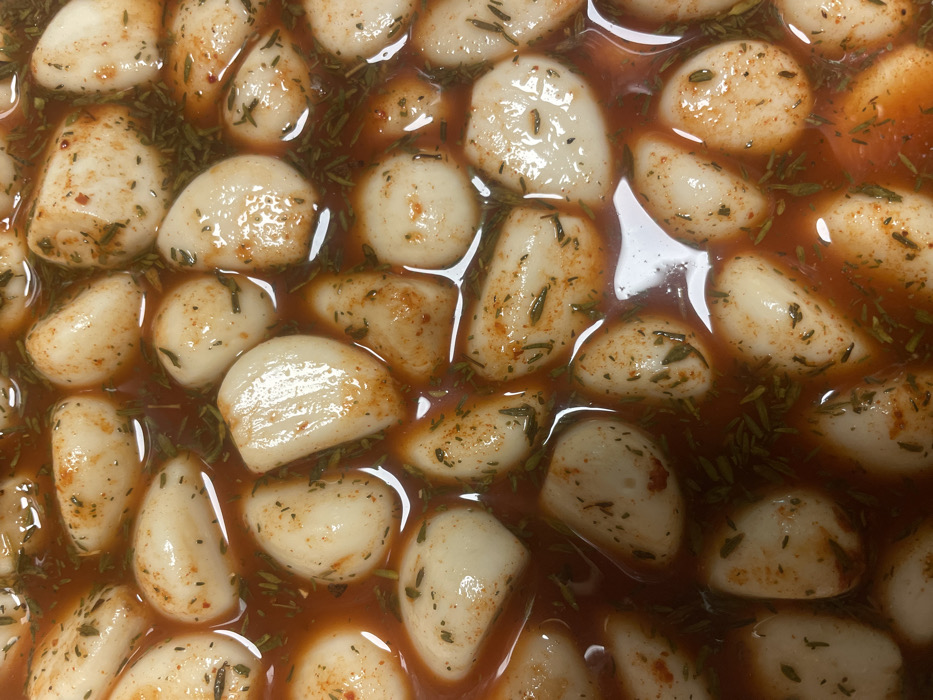 A close-up showcases numerous garlic cloves immersed in a reddish-brown liquid. The garlic cloves are peeled, appearing plump and glossy. They are interspersed with green herbs and flecks of reddish spices, likely chili flakes. The liquid medium reflects light, enhancing the glossy effect on the garlic. The overall composition suggests pickled or preserved garlic.