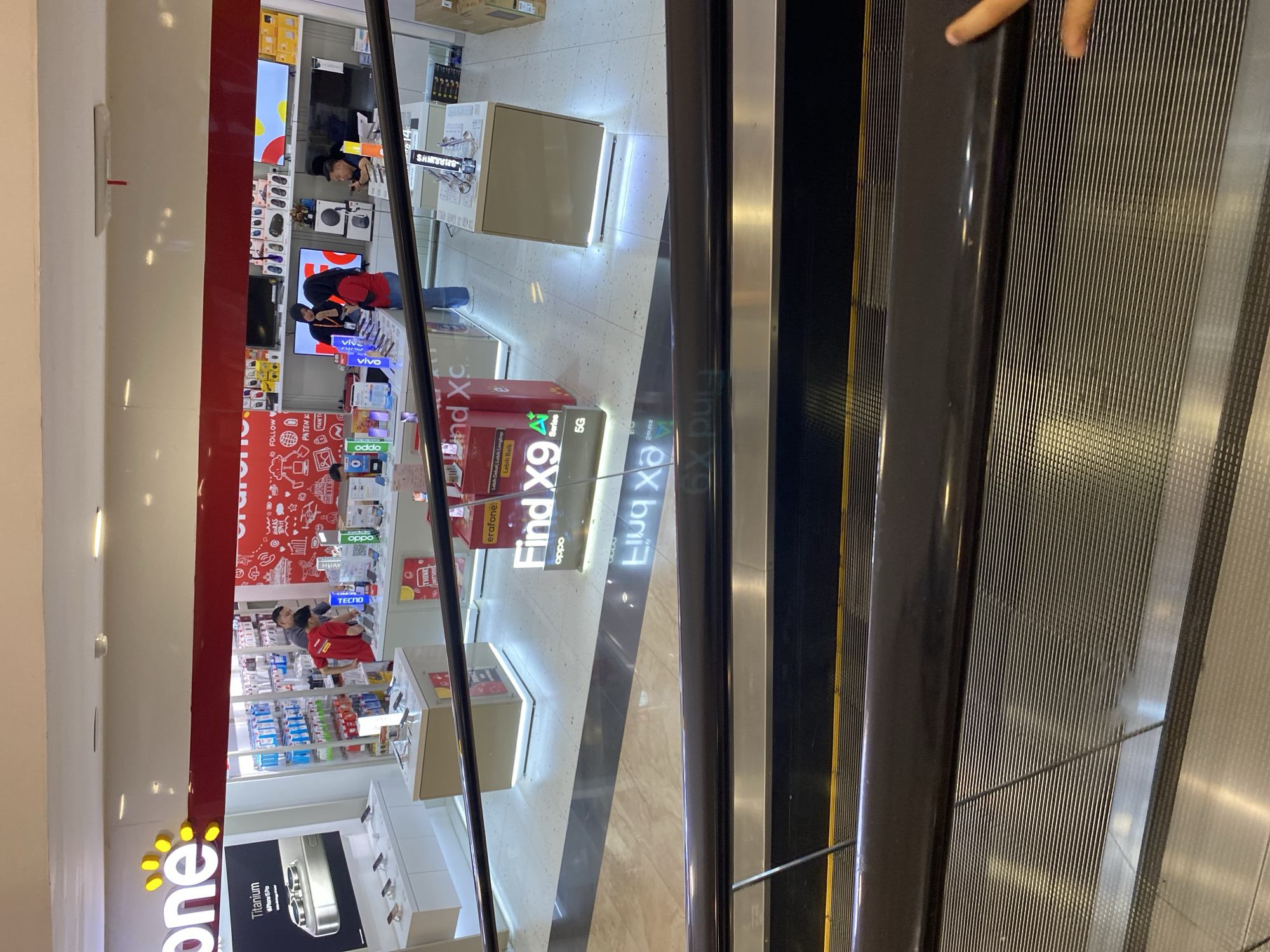 The image depicts the interior of a mobile phone store, likely a franchise given the branding "fone" visible at the top left. The store is well-lit, with displays of mobile phones and accessories. Several individuals are present, appearing to be staff members interacting with customers or organizing merchandise. One person in a red shirt is visible behind a counter, possibly assisting a customer. Another individual in a black hoodie is interacting with a customer, and a third person in a black shirt is seated at a counter. In the foreground, an escalator is partially visible, suggesting the store is located in a mall or a similar multi-level commercial space. The store features promotional signage, including one that reads "Find X9" with the OPPO logo. The overall environment suggests a retail setting focused on consumer electronics, specifically mobile phones. The location is identified as Banda Aceh, Indonesia.