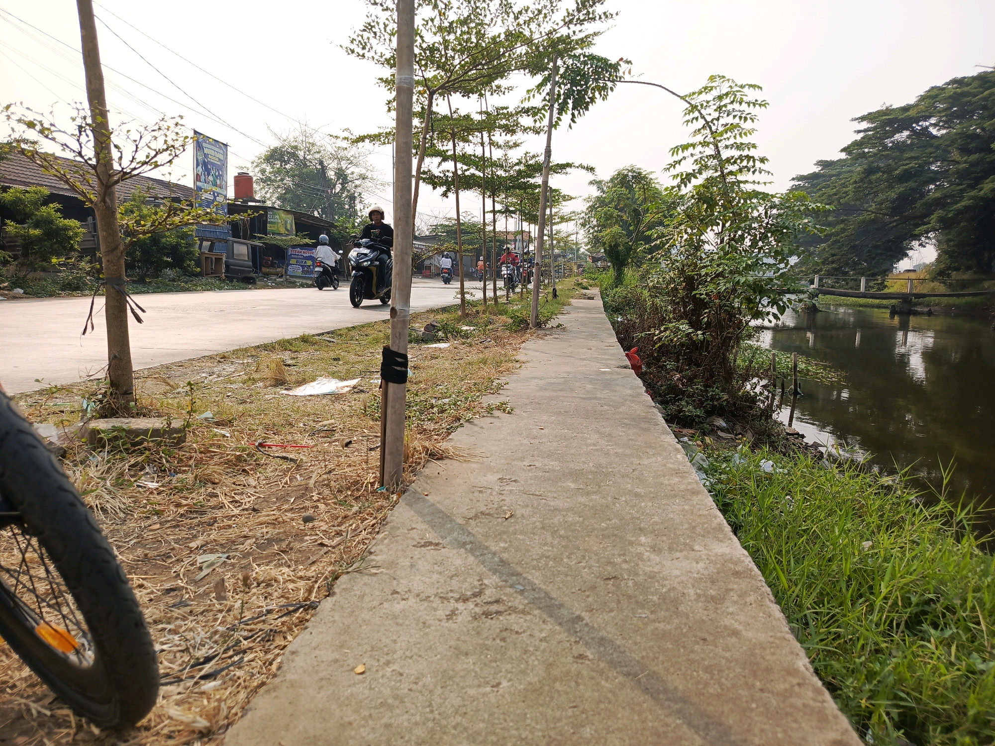 The image depicts a street scene in Tangerang, Indonesia, alongside a canal. The foreground features the edge of a concrete walkway and a bicycle wheel, with dry grass and debris on the left. A row of young trees, staked to vertical poles, lines the edge of the walkway, casting shadows. Beyond the trees, a paved road is visible with several motorcycles in motion, some carrying riders. On the left side of the road, there are small buildings and businesses, including one with a banner displaying Indonesian text and an orange water tank. The right side of the image shows a dark canal with reflections on its surface, bordered by dense green grass and trees. In the distance, a wooden bridge crosses the canal, and lush trees form a canopy over the far bank. The sky is overcast with a pale hue.