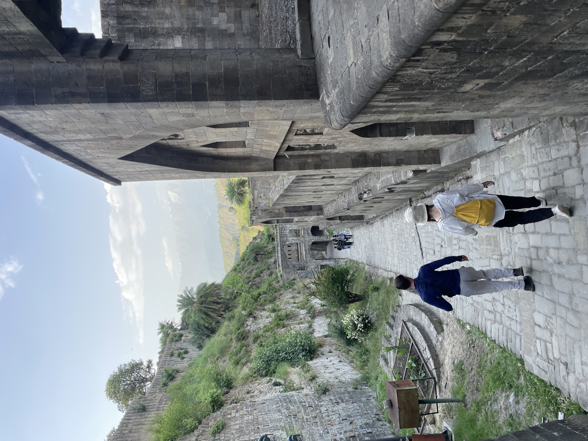 Two individuals walk along a stone-paved pathway between tall stone structures, possibly a fortress or ancient architecture. The path is located in Kaswara, India. The individual closer to the viewer wears a blue long-sleeved shirt and light gray pants. The other individual, further down the path, wears a white shirt with a yellow backpack and black pants. Vegetation grows on the sides of the path. In the distance, beyond the structures, a landscape of rolling hills and trees is visible under a partly cloudy sky.