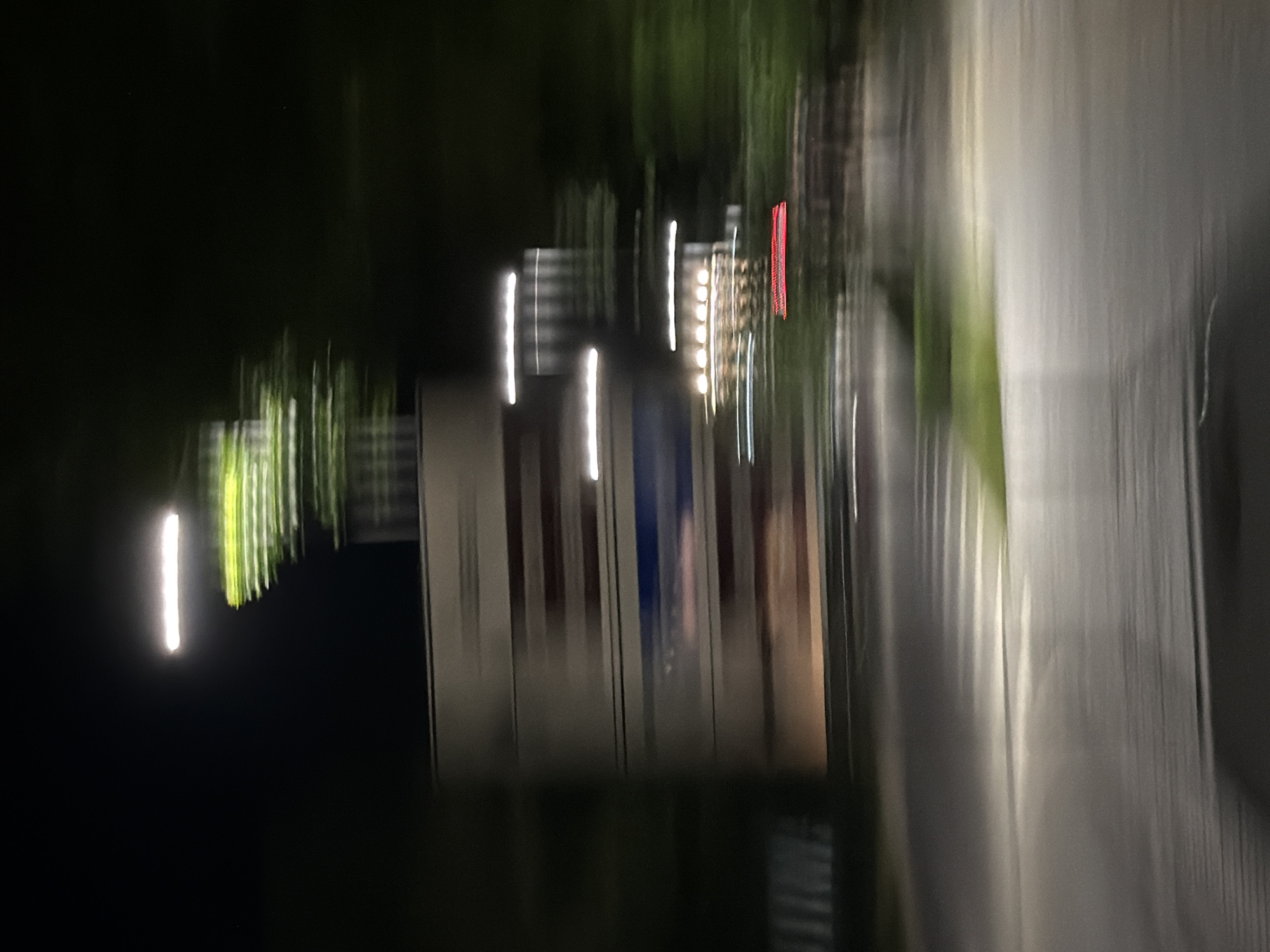 The image captures a motion-blurred nighttime scene in the city of Pravets, Bulgaria. A large, horizontally oriented sign with horizontal stripes, possibly advertising, is visible on the left side of the frame. Above the sign, blurred green foliage and a bright horizontal light suggest street lighting or illumination from the structure. To the right of the sign, a series of horizontal white lights are streaked across the frame, and lower down, a cluster of circular yellow lights and a red rectangular light are also visible, indicative of building lights or signage. The foreground shows a blurred road surface with white lines and patches of green grass, suggesting the image was taken from a moving vehicle. The overall impression is that of a drive-by view at night, with the motion blur obscuring fine details but highlighting the illuminated elements of the urban landscape.