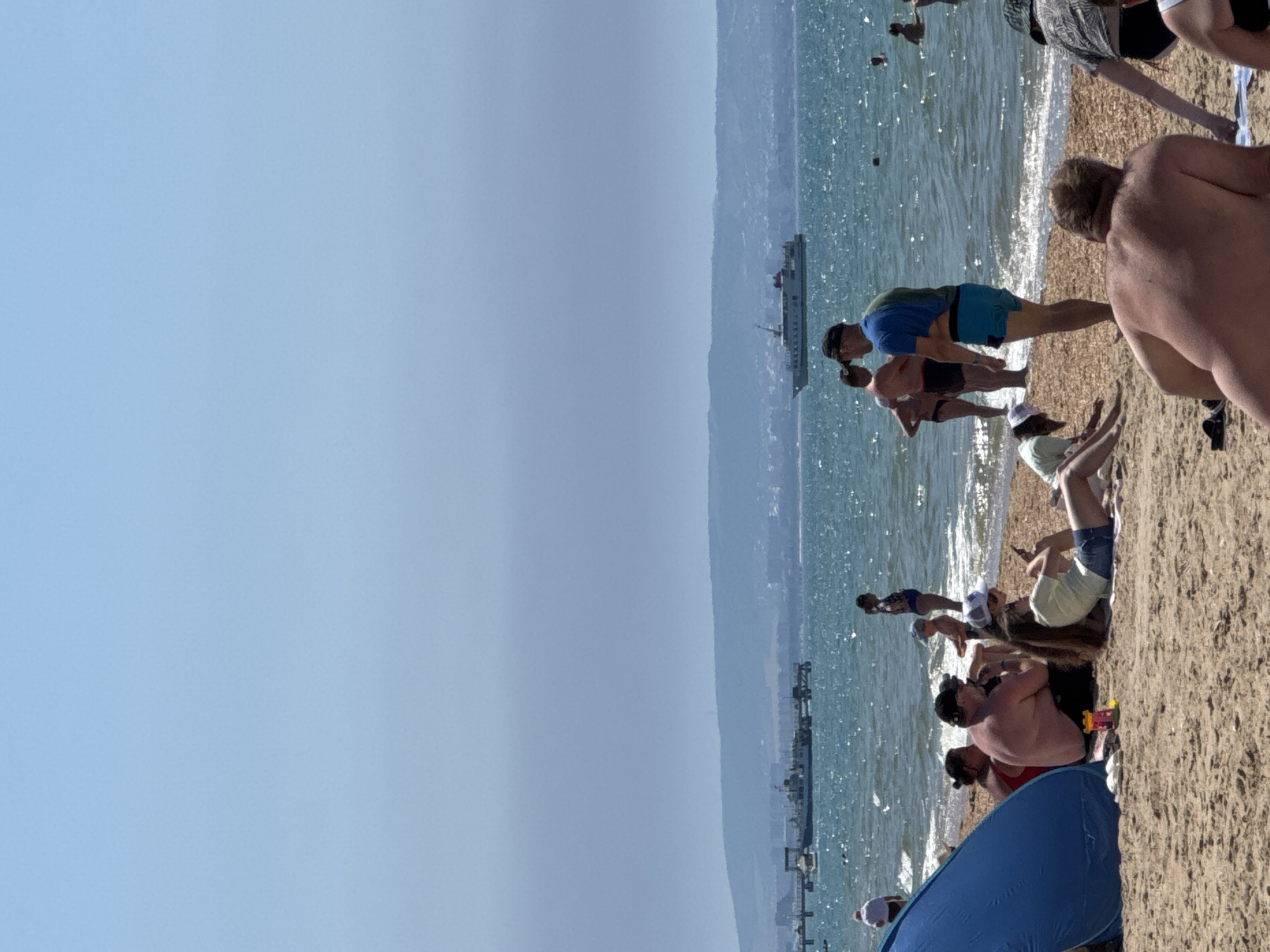 The image depicts a sunny day at a crowded beach in Vityazevo, Russia. The scene is dominated by a sandy shoreline with a clear, blue sky above. The sea, sparkling under the sun, is a prominent feature, with gentle waves lapping at the shore. A large ferry or ship is visible in the distance, sailing on the calm water.
Several groups of people are scattered across the beach, engaged in various activities. Some are sunbathing on towels and blankets, while others are wading in the shallow water. A few individuals appear to be exercising or stretching. The atmosphere suggests a leisurely and enjoyable day at the beach, with a sense of relaxation and recreation.
The people are dressed in swimwear, appropriate for a warm day. The time of day appears to be midday or early afternoon, given the bright sunlight and clear sky. There are no visible signs or text in the image. The overall impression is one of a typical summer day at a popular seaside resort.