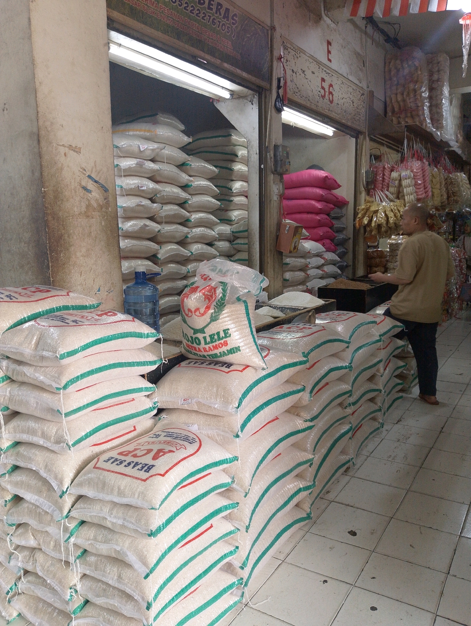 A man in a tan shirt and dark pants stands to the right of a stall piled high with bags of rice. The stall appears to be in a market in Lembang, Indonesia, indicated by signage that reads "BERAS" (rice) in large letters at the top of the stall and a stall number "56" above an entrance. The man is facing away from the camera, reaching into a dark wooden bin that contains loose grain. In the foreground, stacks of white rice bags with green and red stripes dominate the scene. A blue water cooler jug sits on the floor between some of the rice bags. Further back, in the entrance of the stall, are stacks of pink and gray sacks. Shelves above and to the right are laden with packages of food items, possibly snacks or dried goods, and strings of hanging merchandise. The floor is tiled.